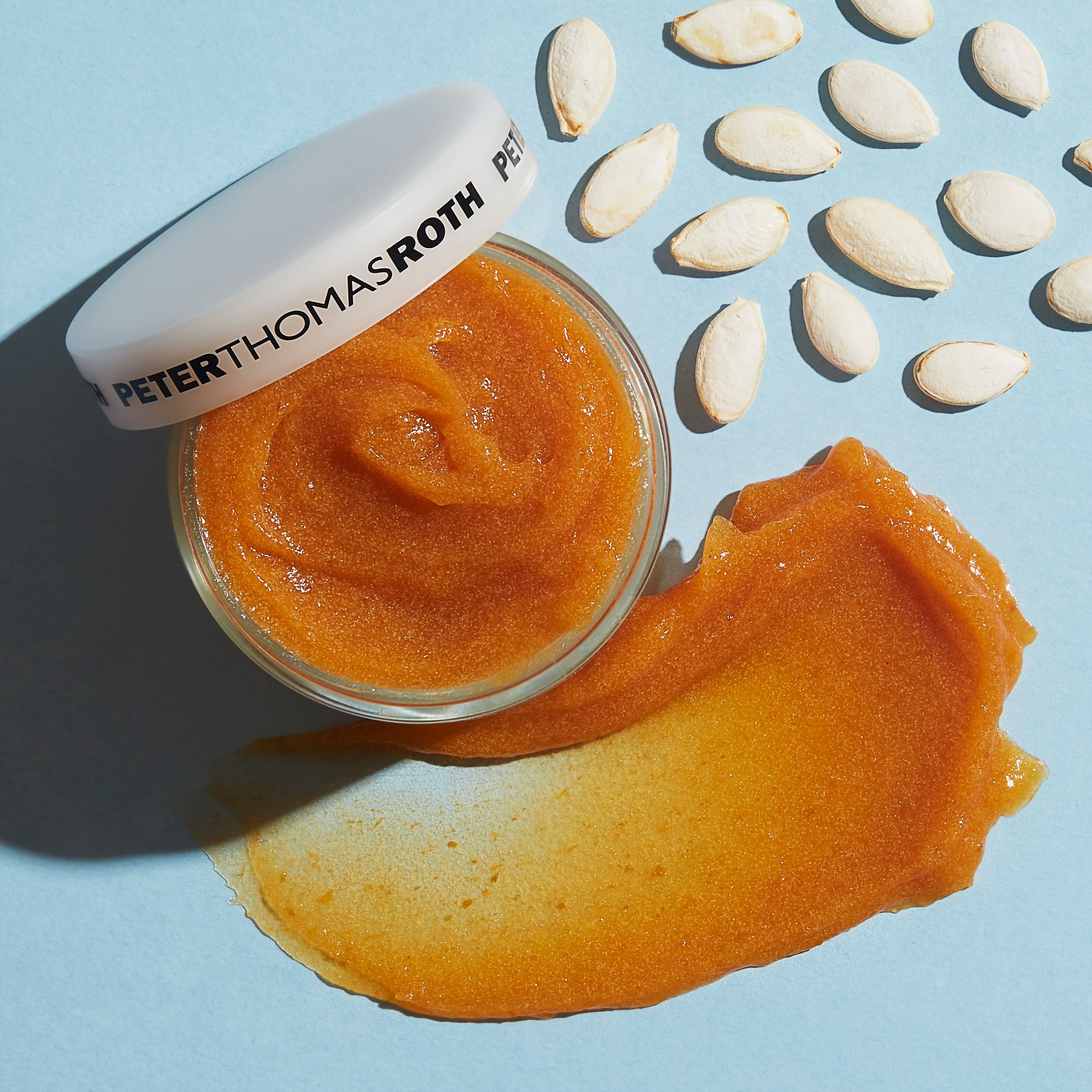 Offenes Tiegel mit orangefarbener Maske und Klecks. Deckel mit Markennamen. Kürbiskerne auf blauem Hintergrund.