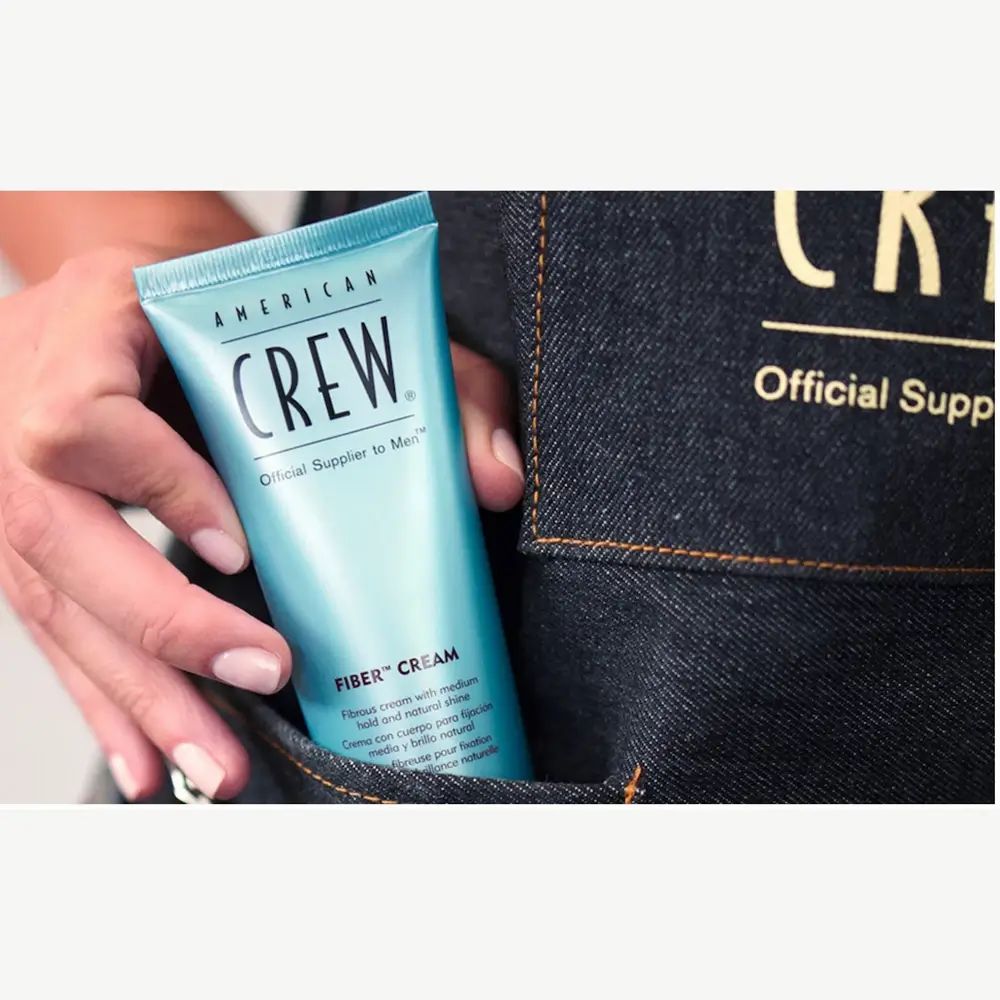 Hellblaue Tube in einer Hand. Aufschrift: American Crew, Fiber Cream. Vor Jeans-Hintergrund.