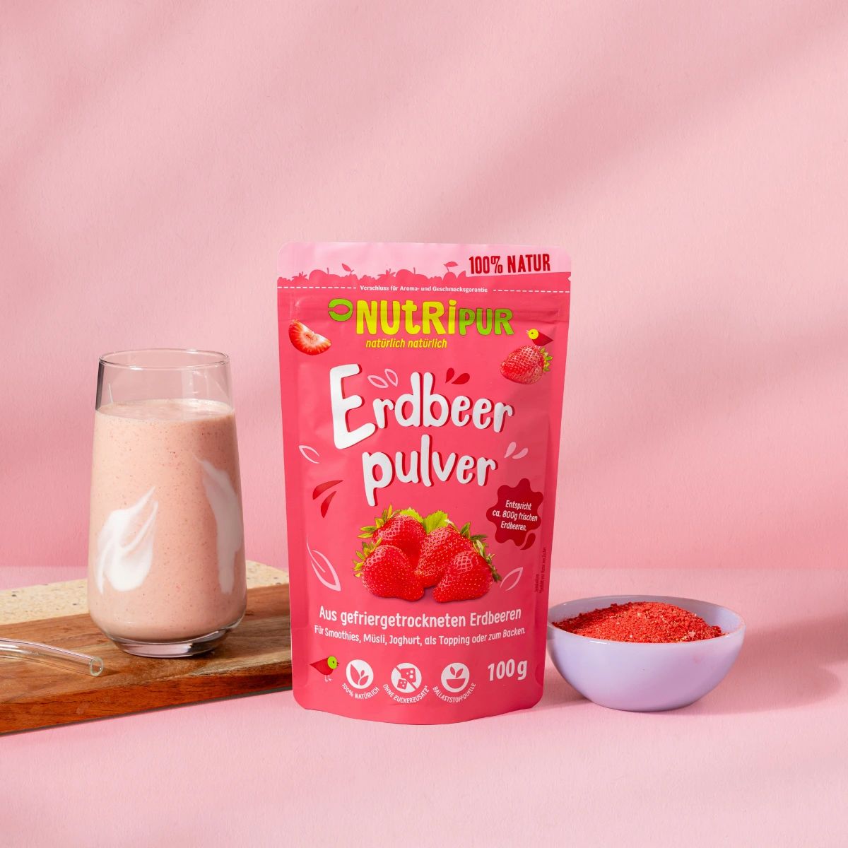 Ein Glas mit rosa Smoothie, daneben ein Beutel Erdbeerpulver und eine Schale mit Pulver.