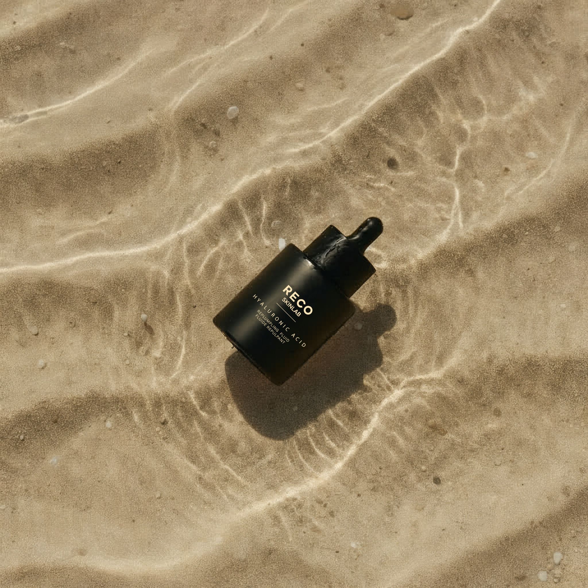 Schwarze Flasche mit Pipette im Wasser. Aufschrift: RECO SKINLAB, HYALURONIC ACID, REPLUMPING FLUID.