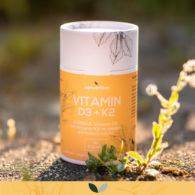 Zylindrische Dose Sanutrition Vitamin D3 + K2 im Freien. Orangefarbene Etikett mit Produktinformationen. Blätter und Pflanze im Vordergrund.
