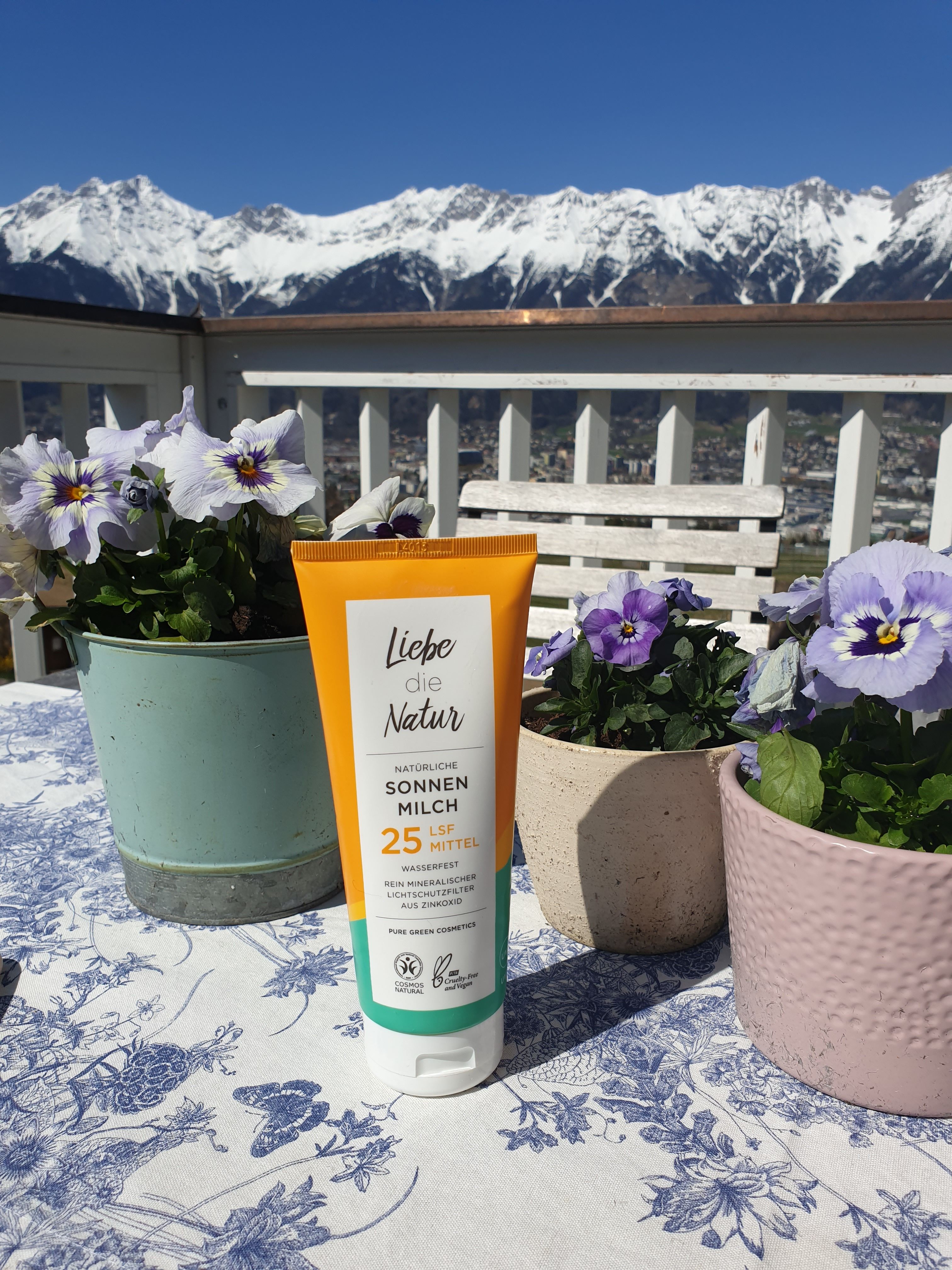 Sonnenmilch-Tube mit gelb-weißem Etikett. Text: Natürliche Sonnenmilch LSF 25 Mittel. Wasserfest. Bio-Siegel. Vor Blumen und Bergpanorama.