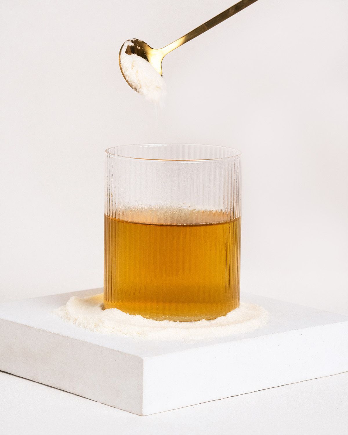 Glas mit Flüssigkeit und Pulver. Ein Löffel hält Pulver über dem Glas. Auf weißem Podest.