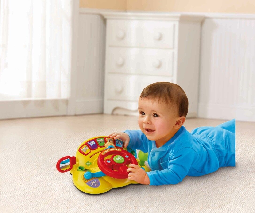Baby liegt auf dem Bauch und spielt mit einem gelben Spielzeug-Lenkrad. Das Kind lächelt.