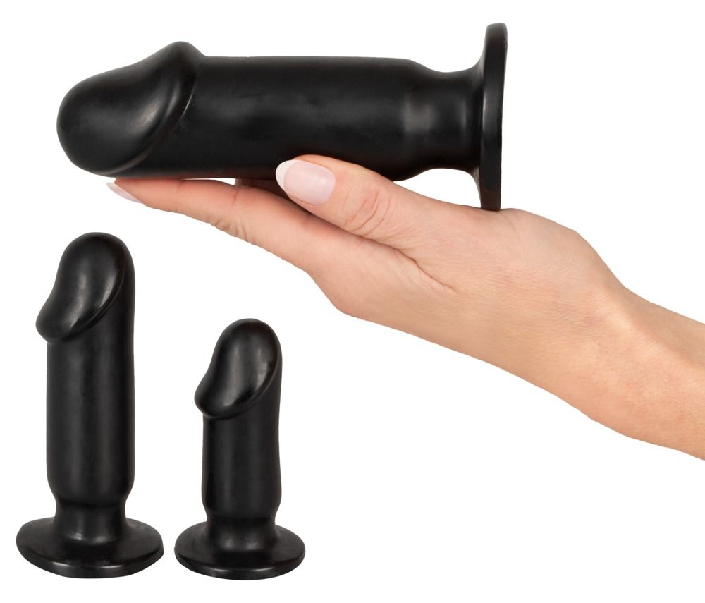 Drei schwarze Analplugs. Ein Plug wird von einer Hand gehalten. Die anderen zwei liegen daneben. Zylindrische Form mit abgerundeter Spitze.