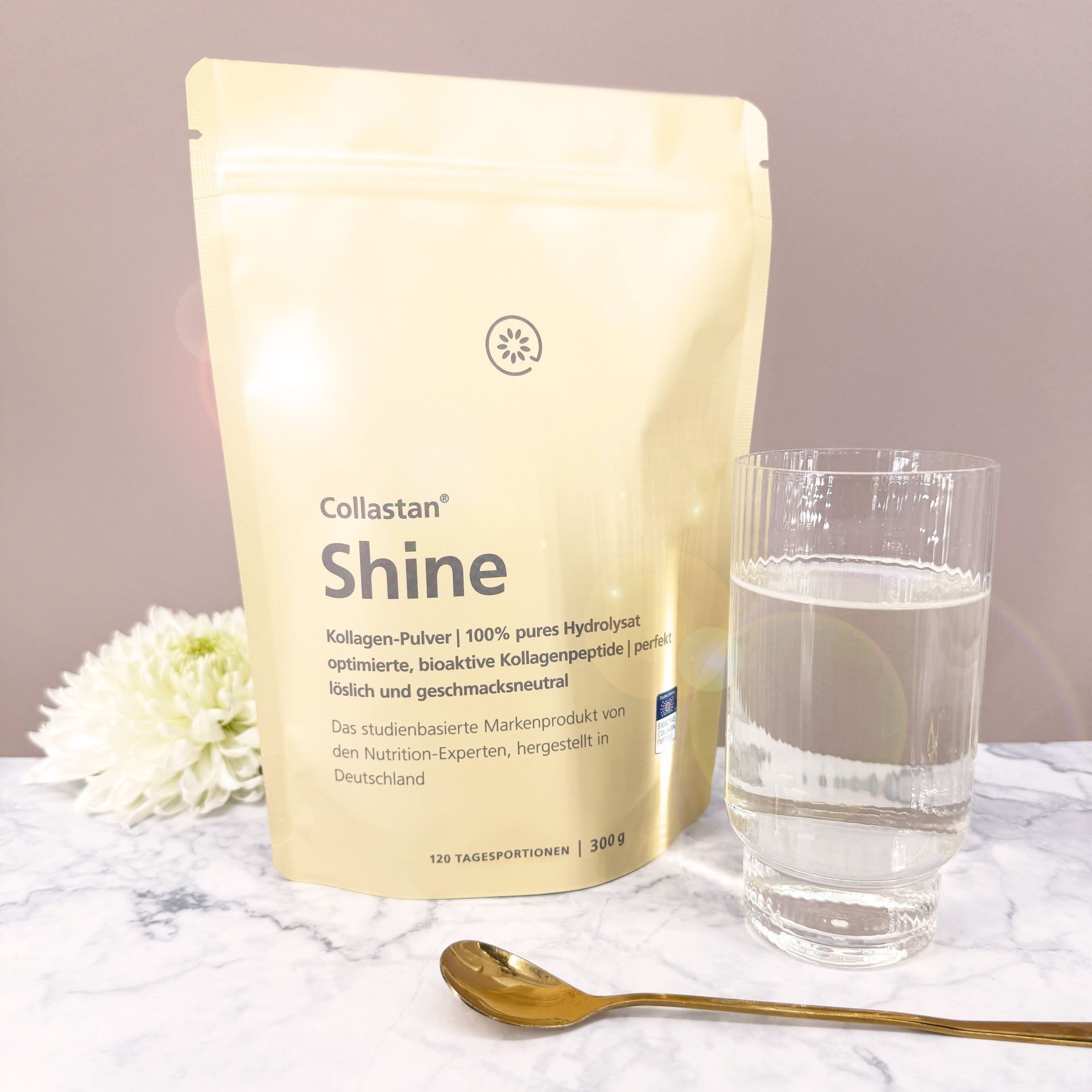 Produktverpackung "Collastan Shine" neben Glas Wasser und Löffel. Hintergrund: Blume.