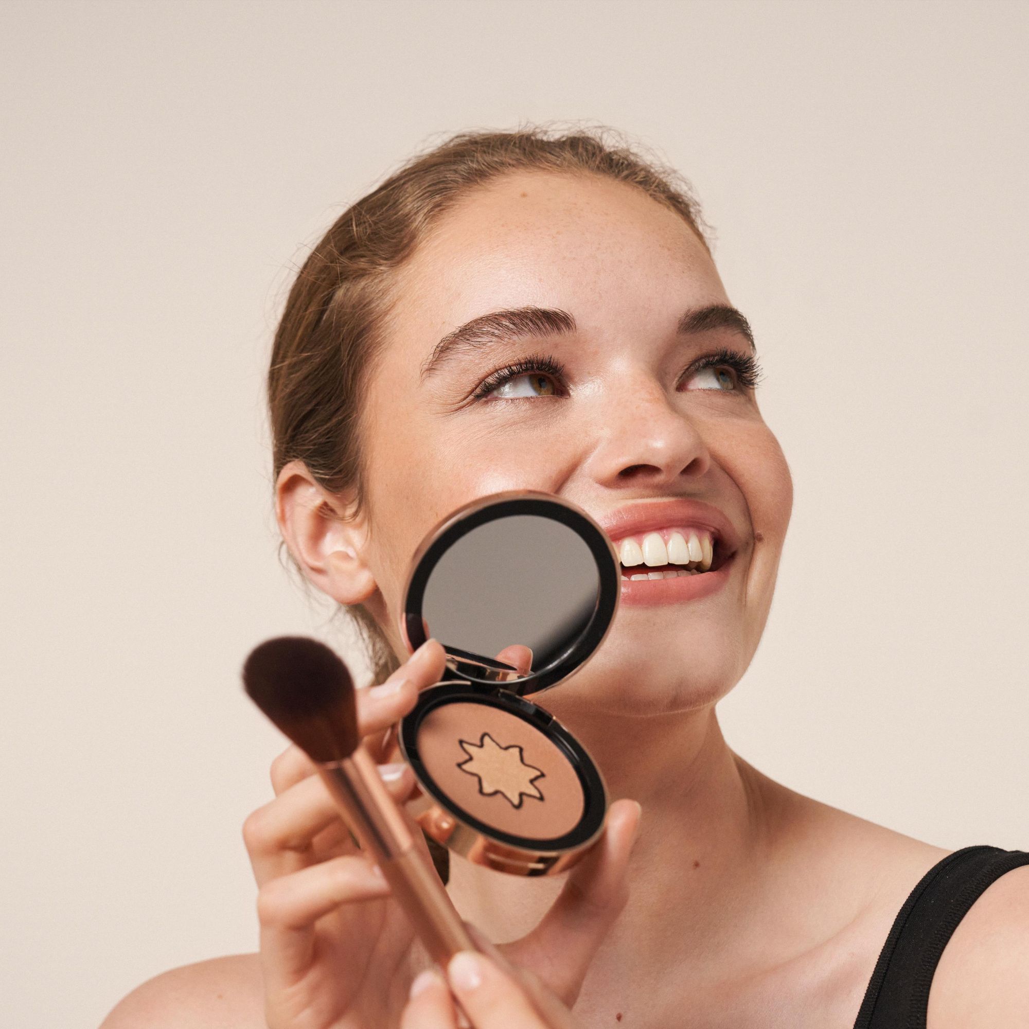 Frau hält Bronzer-Dose und Pinsel. Sie lächelt. Dose geöffnet, Spiegel sichtbar.