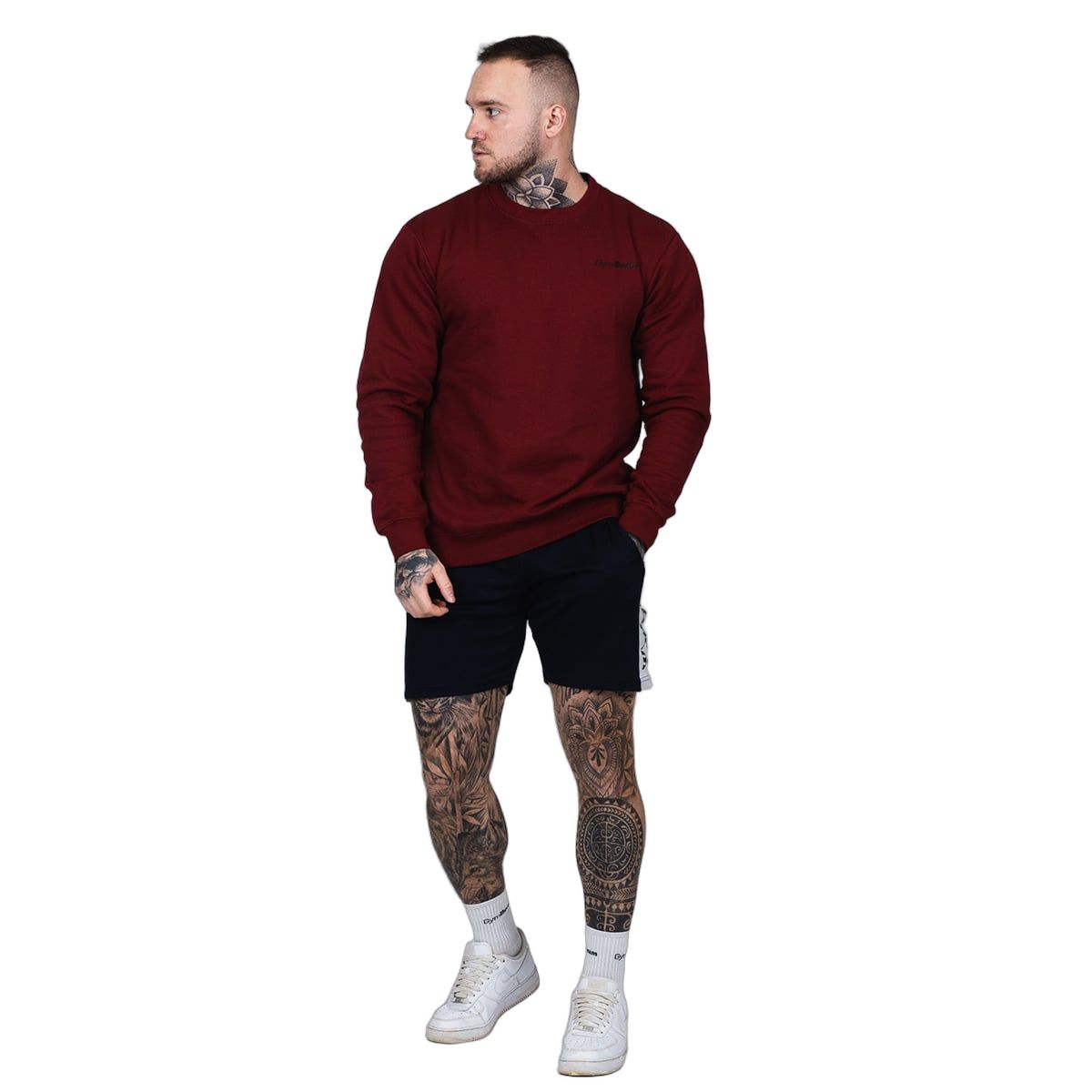 Mann mit Tätowierungen steht in einem burgunderroten Pullover, schwarzen Shorts und weißen Sneakern.