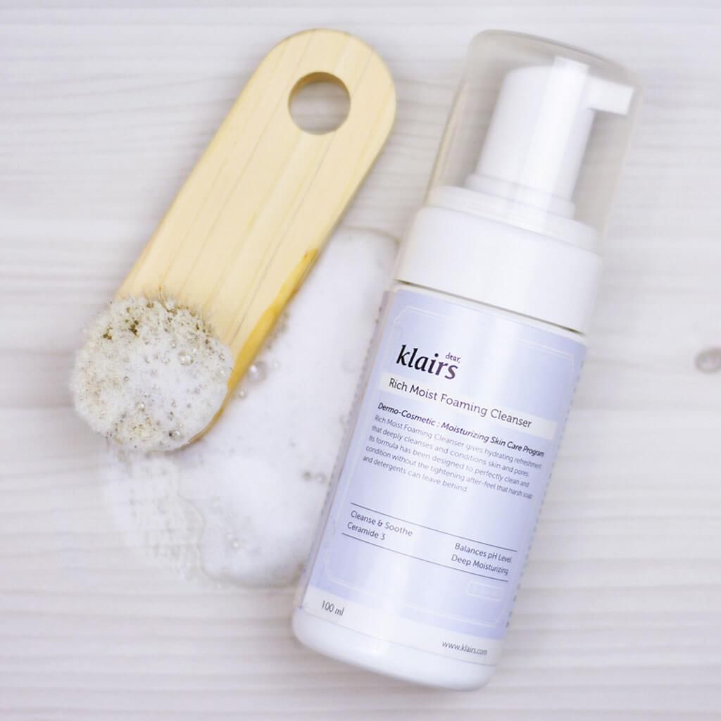 Weiße Flasche Klairs Rich Moist Foaming Cleanser mit Bürste. Schaum daneben. Holzbrett. Blaue Etikett mit Text.