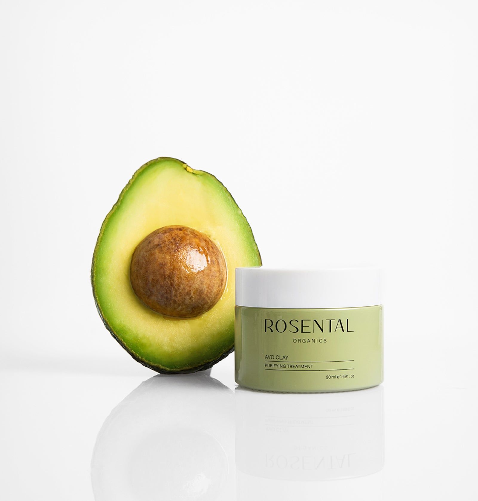 Halbe Avocado neben einem Rosental Avo Clay Mask-Glas. Weißer Hintergrund. Produkt und Frucht sind reflektiert.