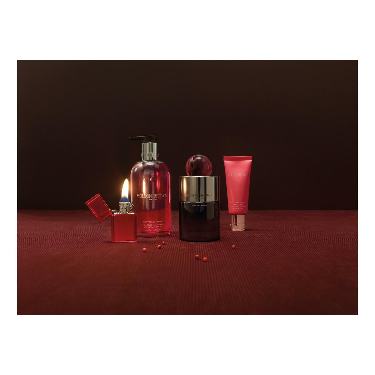 Produkt-Arrangement auf rotem Untergrund. Enthält Flasche, Parfum, Creme, Feuerzeug und Beeren. Produkte sind rot.