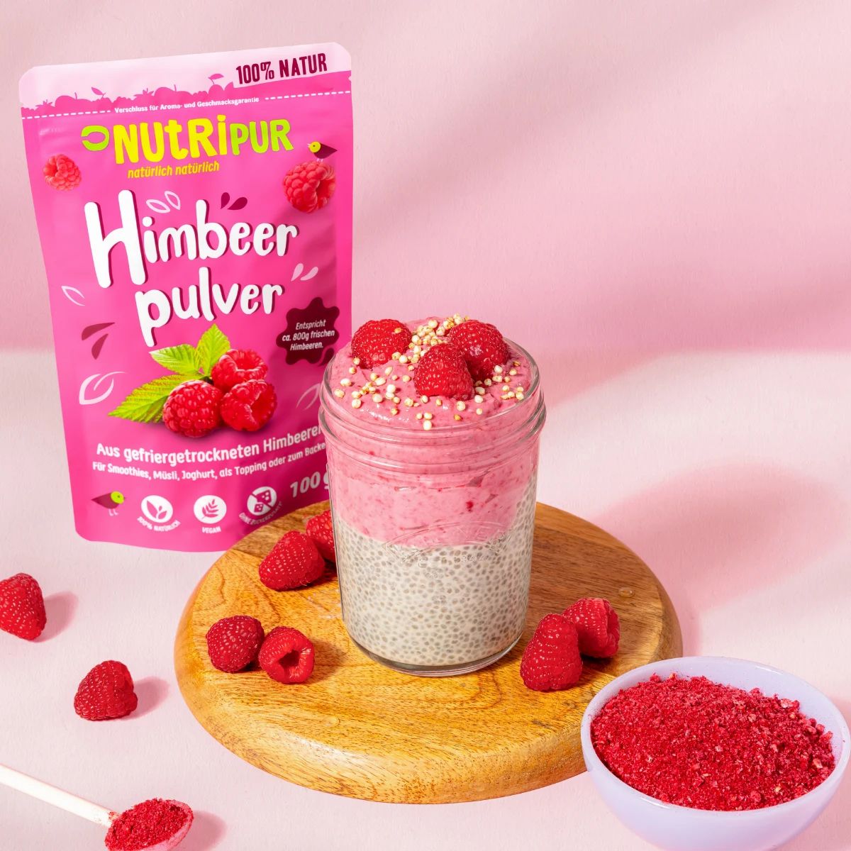 Himbeerpulver in einem Glas mit Chia-Pudding und frischen Himbeeren. Daneben eine Packung Himbeerpulver und eine Schale mit Pulver.