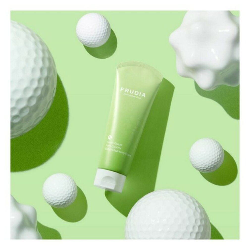 Grüne Tube mit weißem Verschluss. Aufschrift: FRUDIA. Text: Green Grape Pore Control Scrub Cleansing Foam. Umgeben von weißen Objekten.