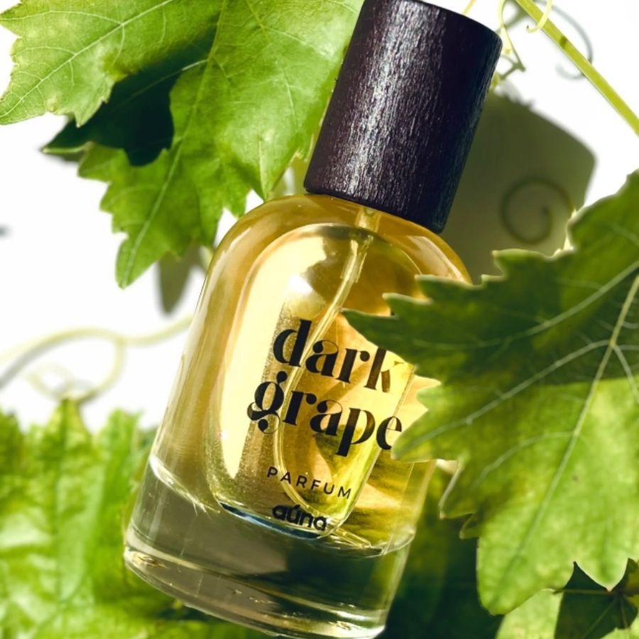 Parfumflasche mit braunem Deckel, umgeben von grünen Blättern. Aufschrift "dark grape parfum" und "auna".