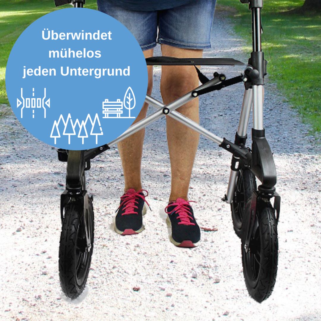 Rollator von vorne. Schwarze Räder, silbernes Gestell. Person mit Schuhen im Bild.