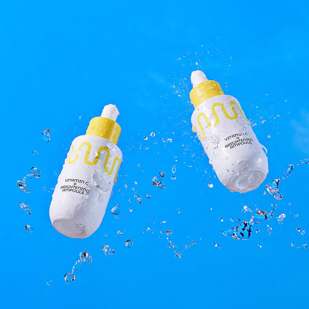 Zwei weiße Pipettenflaschen mit gelben Verschlüssen, umgeben von Wasser. Enthält Vitamin C Brightening Ampoule.