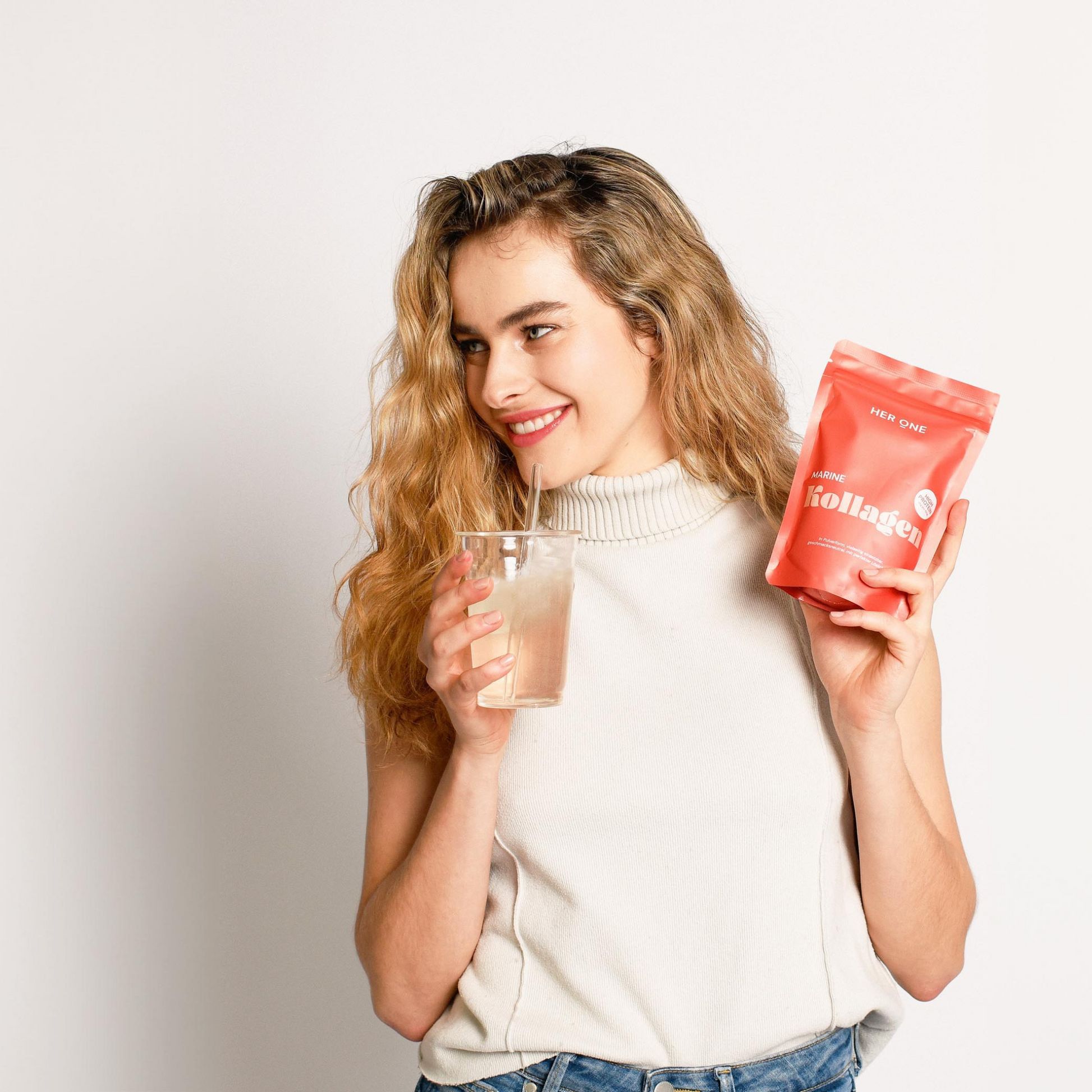 Frau hält ein Glas Getränk und eine rote Tüte mit der Aufschrift "HER ONE Kollagen".