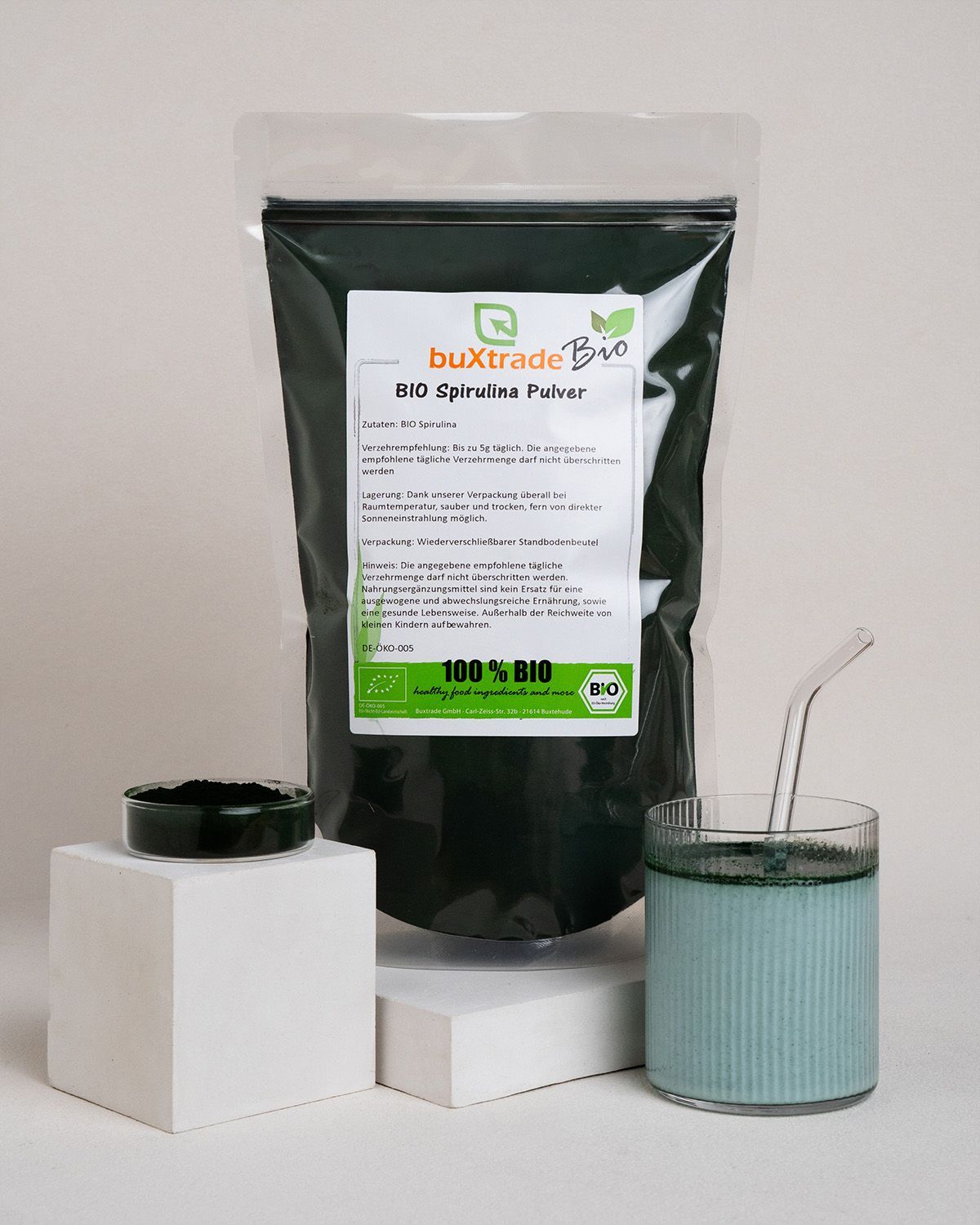 Glas mit blauem Getränk und Spirulina-Pulver. Kleines Schälchen mit Pulver. Transparente Plastikverpackung mit Produkt.
