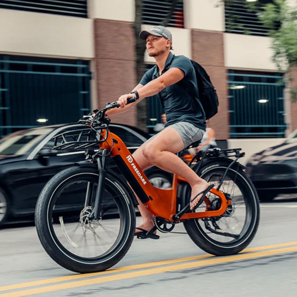 Mann fährt orangefarbenes Elektrofahrrad auf einer Straße. Marke FREEDARE. Schwarze Reifen, Gepäckträger.