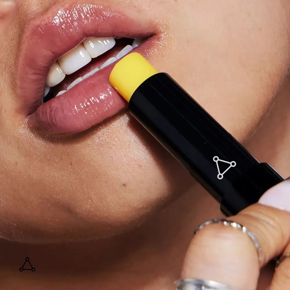 Person benutzt einen schwarzen Lippenbalsam-Stick mit gelber Spitze. Lippen sind sichtbar.