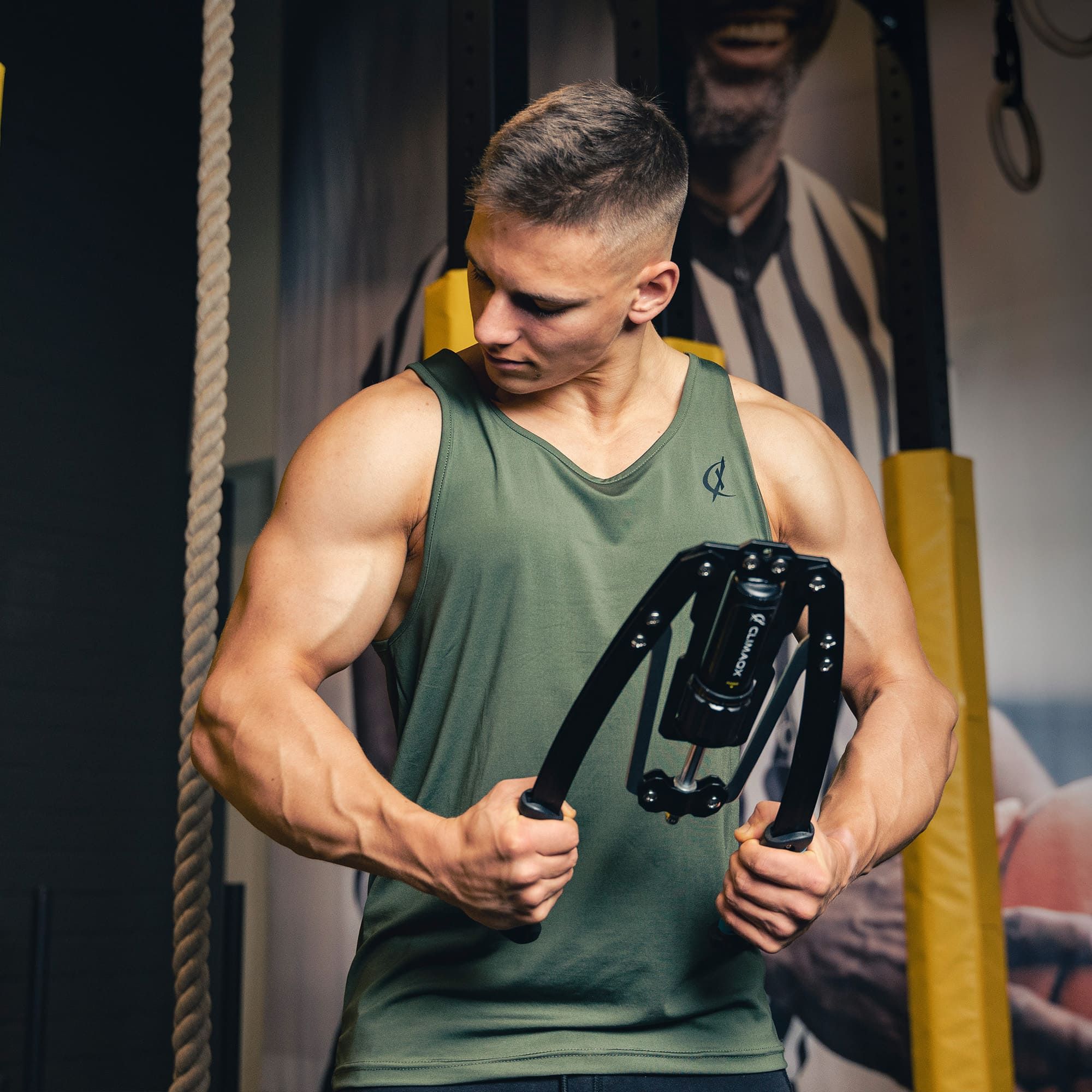 Mann hält ein schwarzes Fitnessgerät. Er trägt ein grünes Tanktop. Im Hintergrund ein Fitnessstudio.