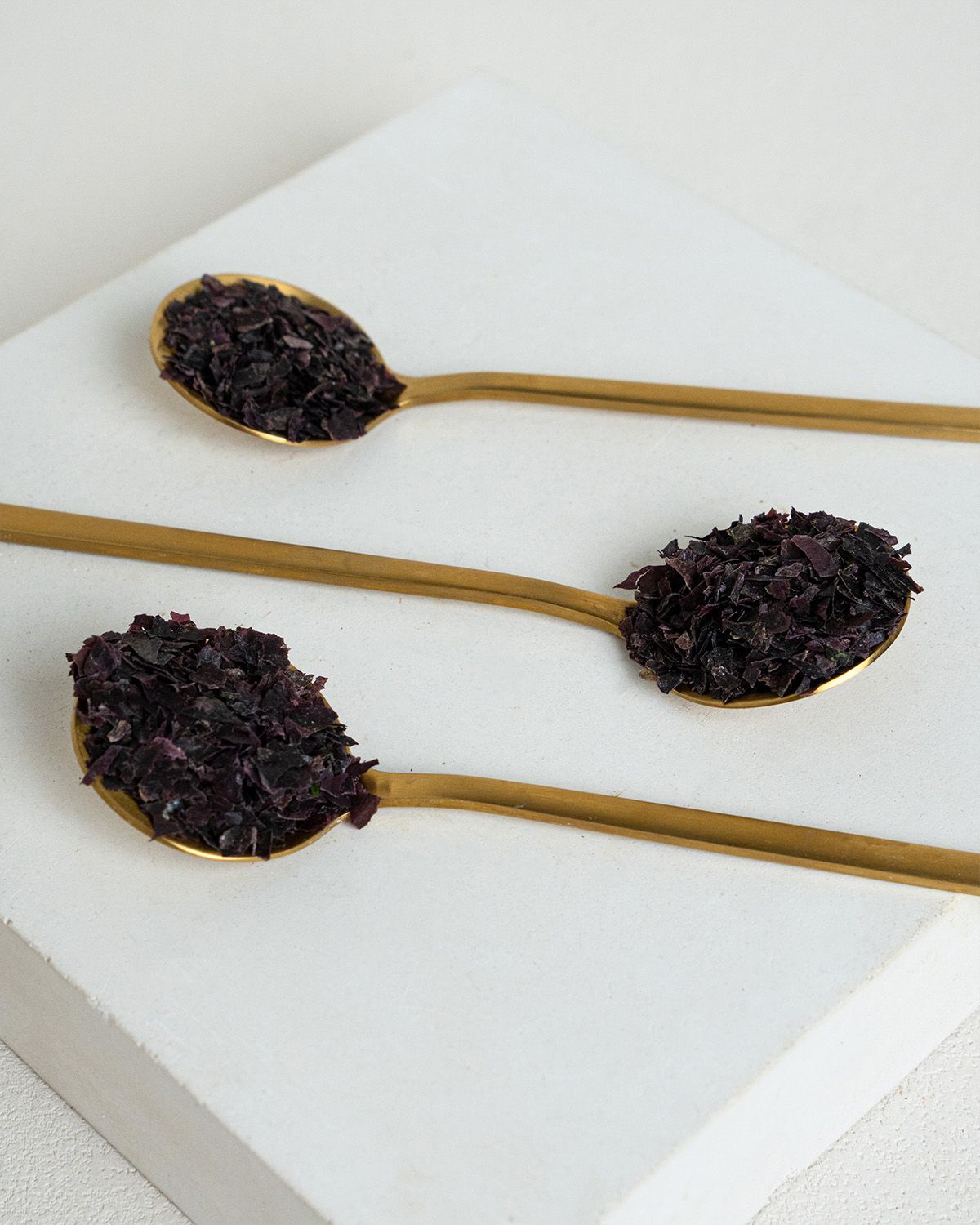Drei goldene Löffel mit Bio Dulse Flocken auf einer weißen Oberfläche. Nahaufnahme der Flocken.