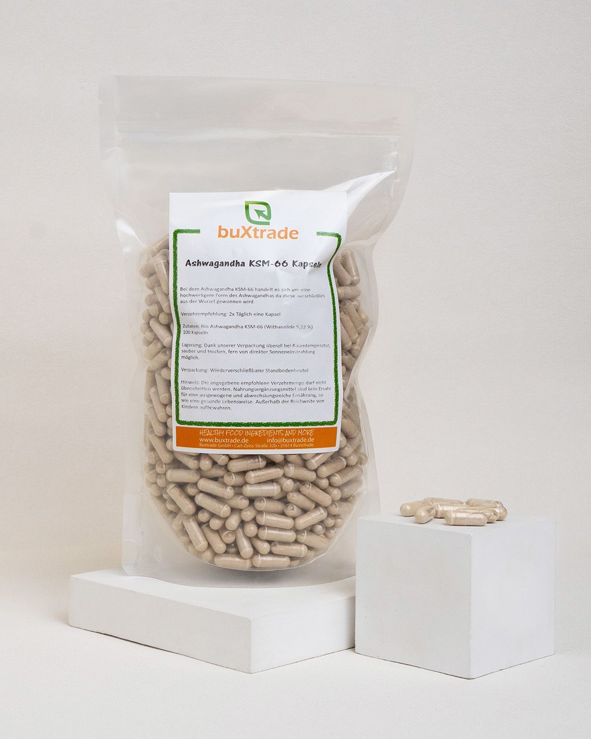Beutel mit Kapseln. Aufdruck: buXtrade Ashwagandha KSM-66 Kapseln. Etikett mit Text. Einige Kapseln daneben.