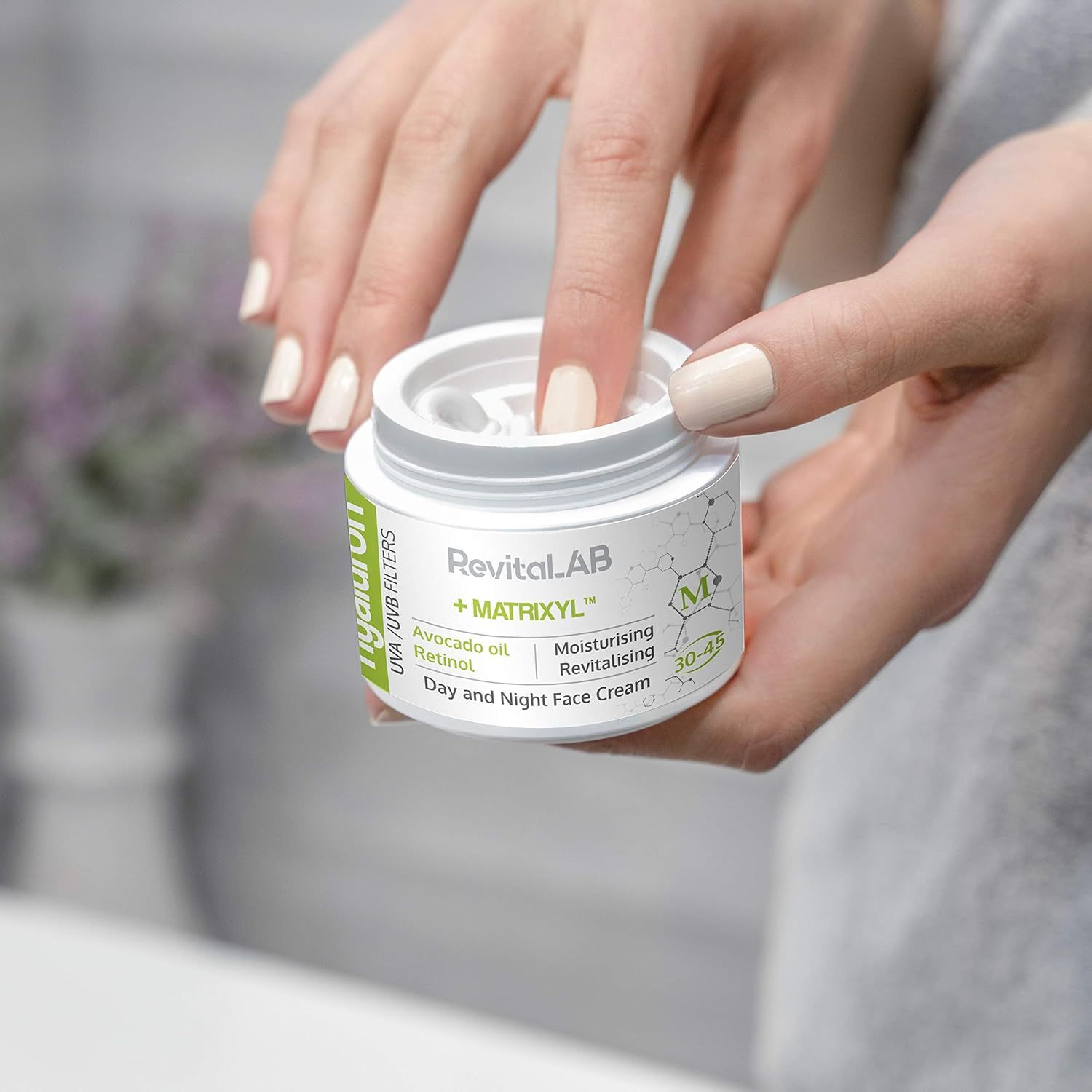 Hände halten einen weißen Tiegel mit Deckel. Aufschrift: RevitaLAB + Matrixyl, Hyaluron, UVA/UVB Filters, Avocadoöl, Retinol, Tages- und Nachtcreme. Altersempfehlung 30-45.