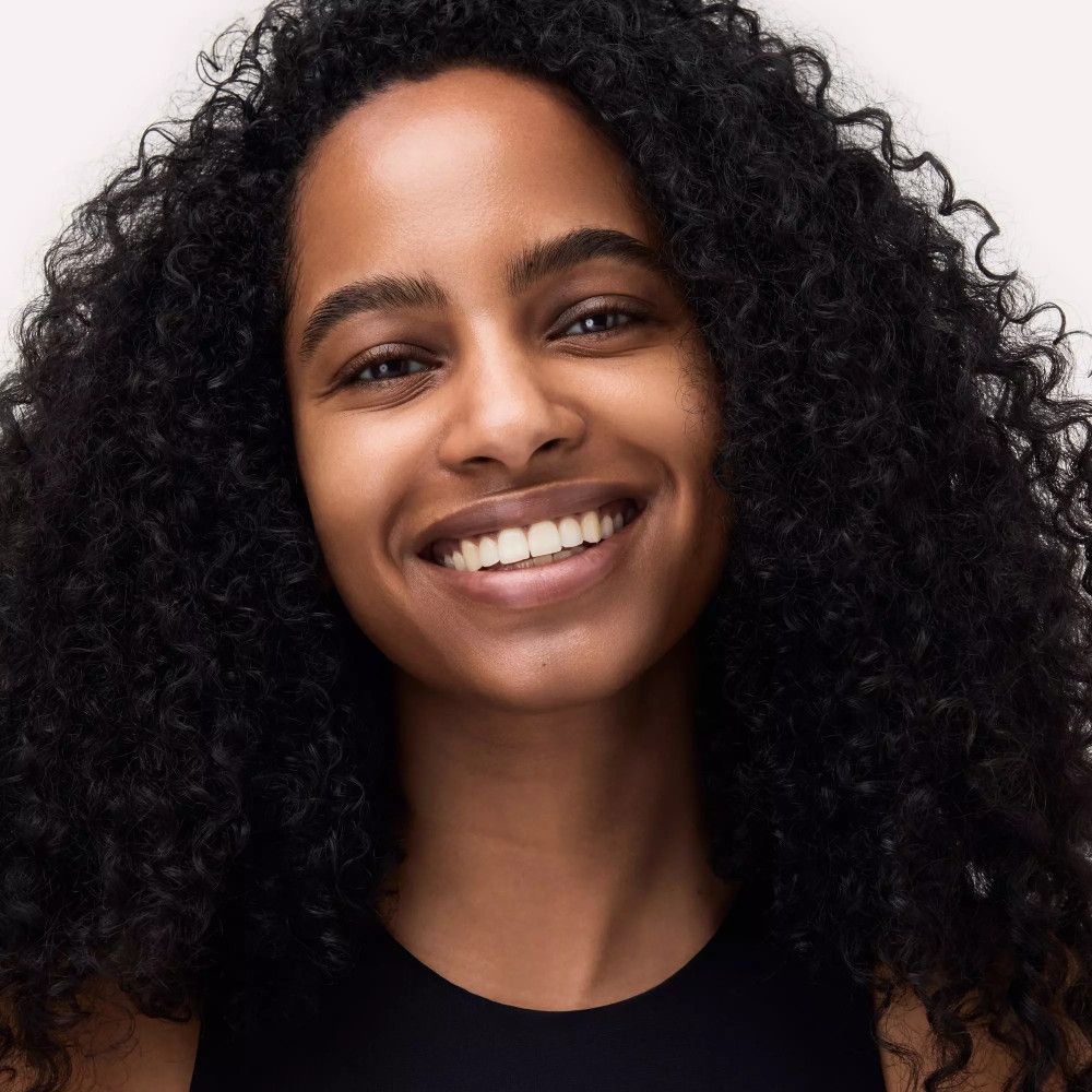 Porträt einer lächelnden Frau mit lockigem, schwarzem Haar. Sie trägt ein schwarzes Top.