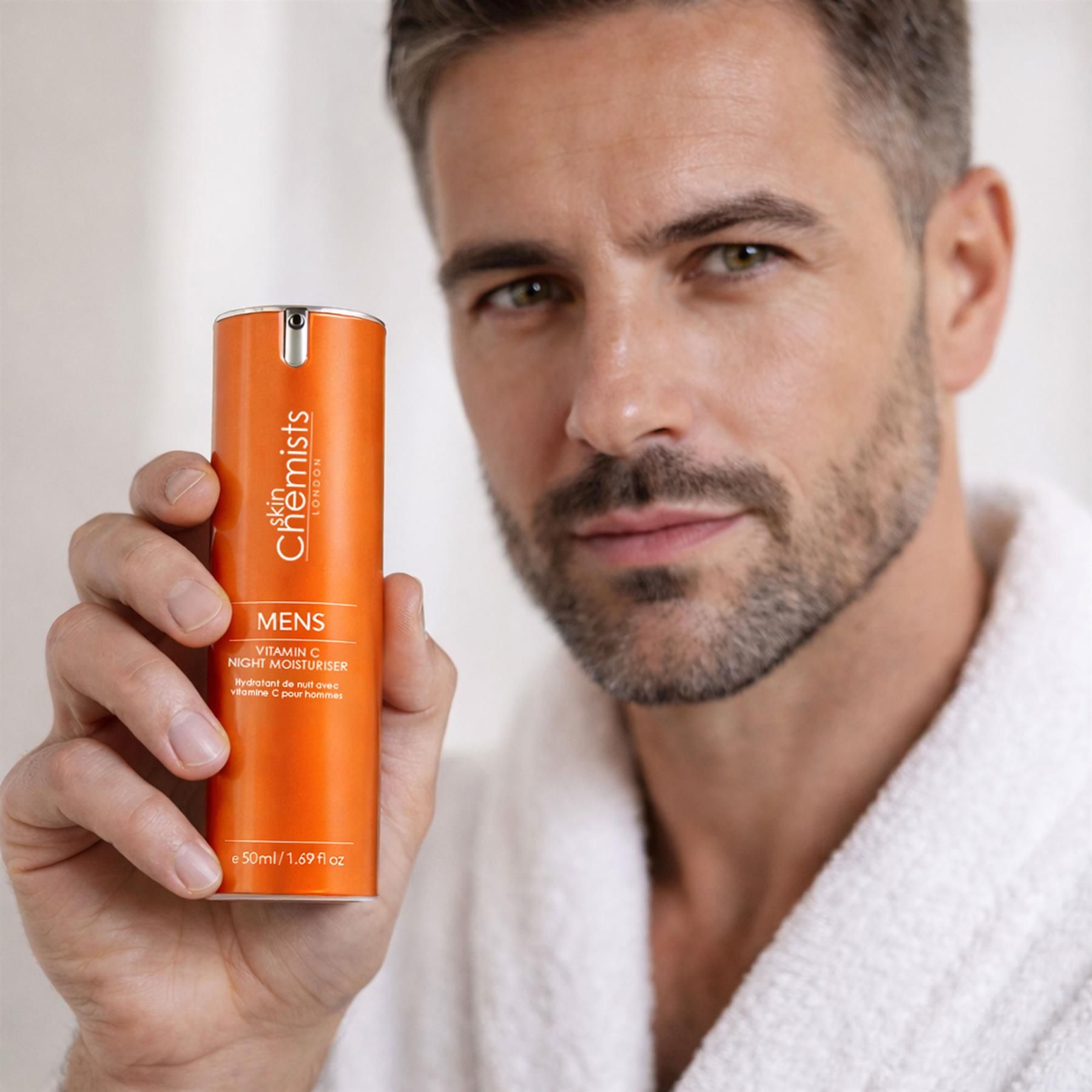 Ein Mann im Bademantel hält eine orangefarbene Flasche SkinChemists Vitamin C Nacht-Feuchtigkeitscreme.