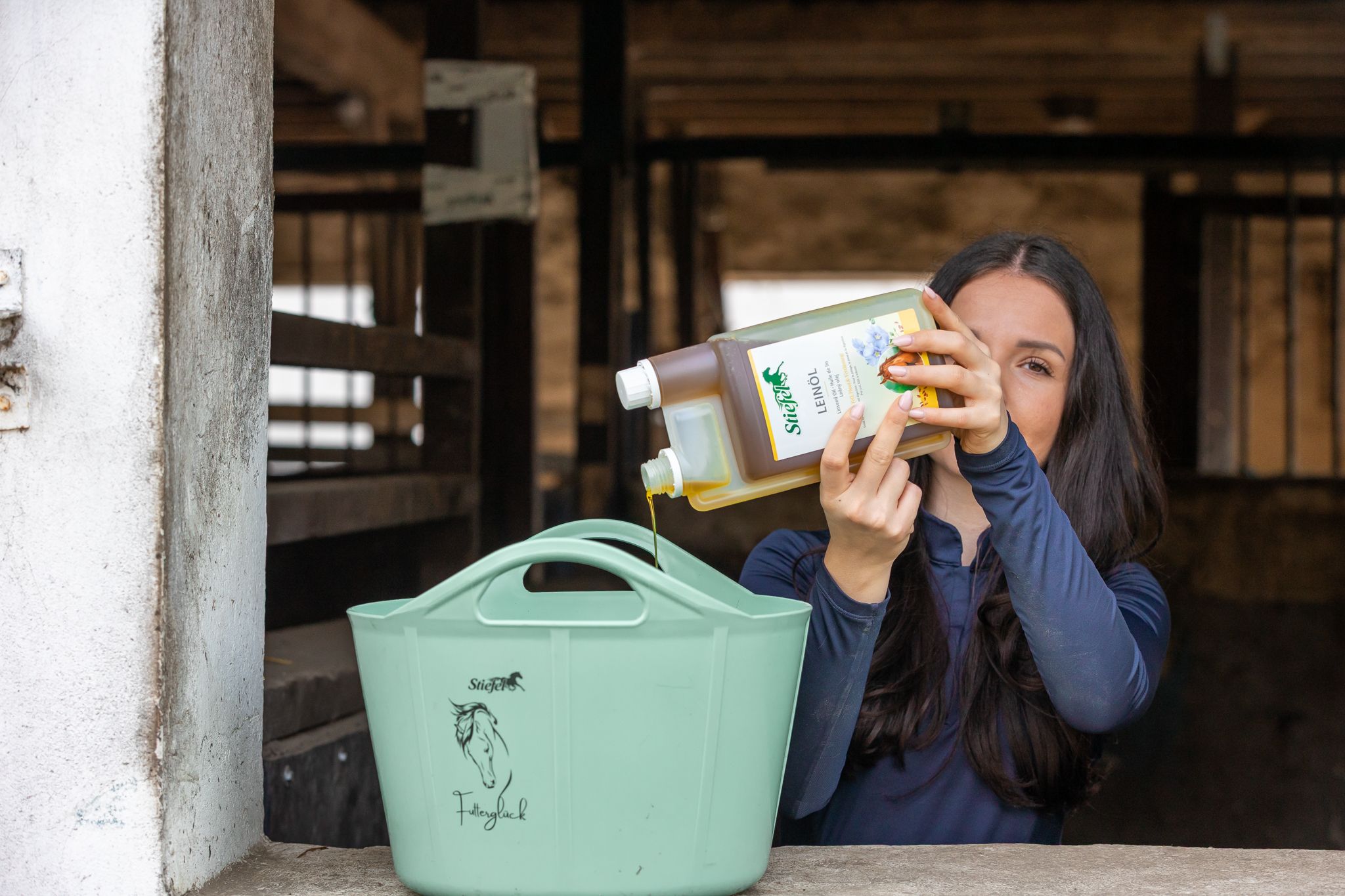 Person gießt Stiefel Leinöl aus einer Flasche in einen grünen Eimer. Hintergrund: Stall.