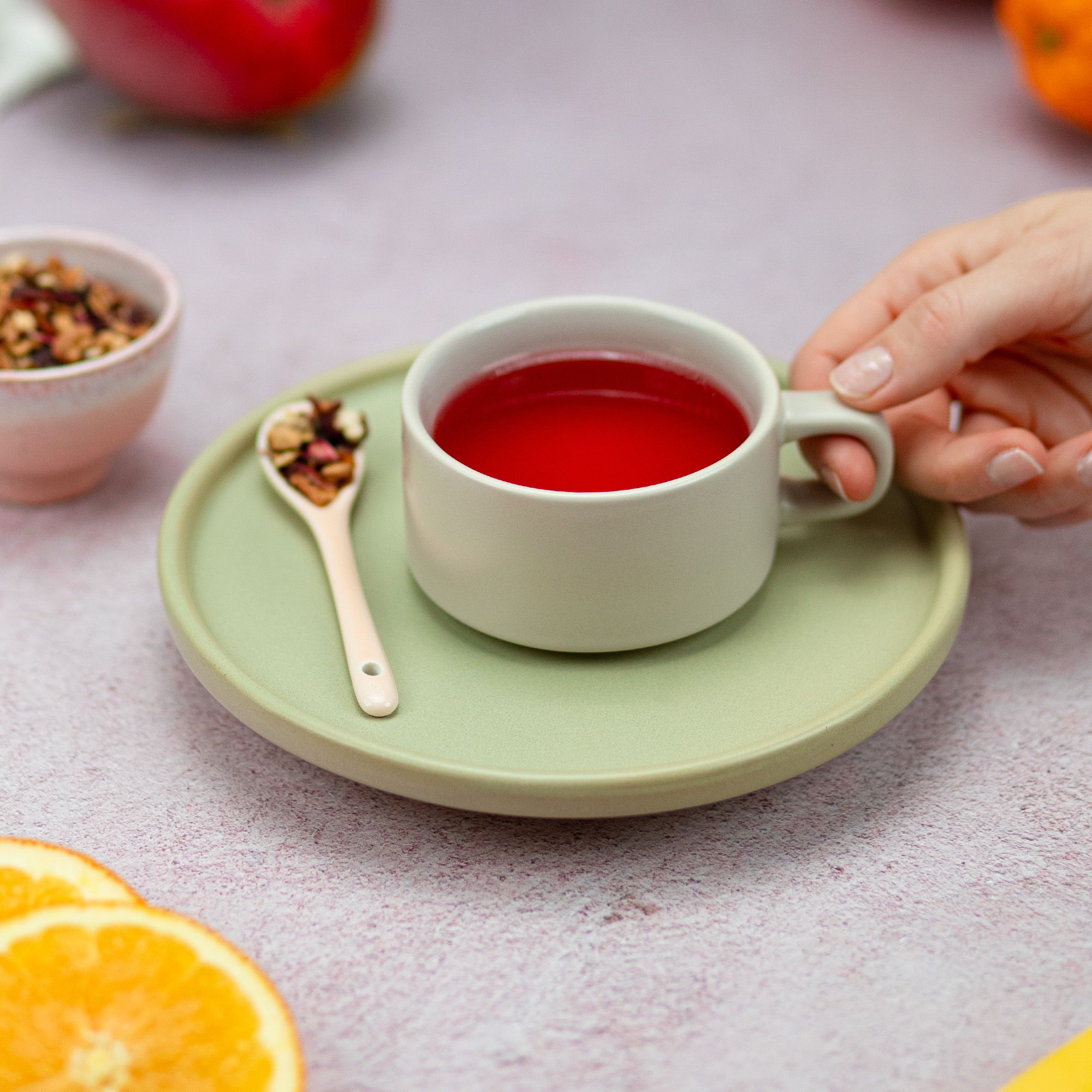 Tasse Tee auf Untertasse. Löffel mit Teemischung daneben. Hand hält die Tasse. Orangenstücke im Hintergrund.