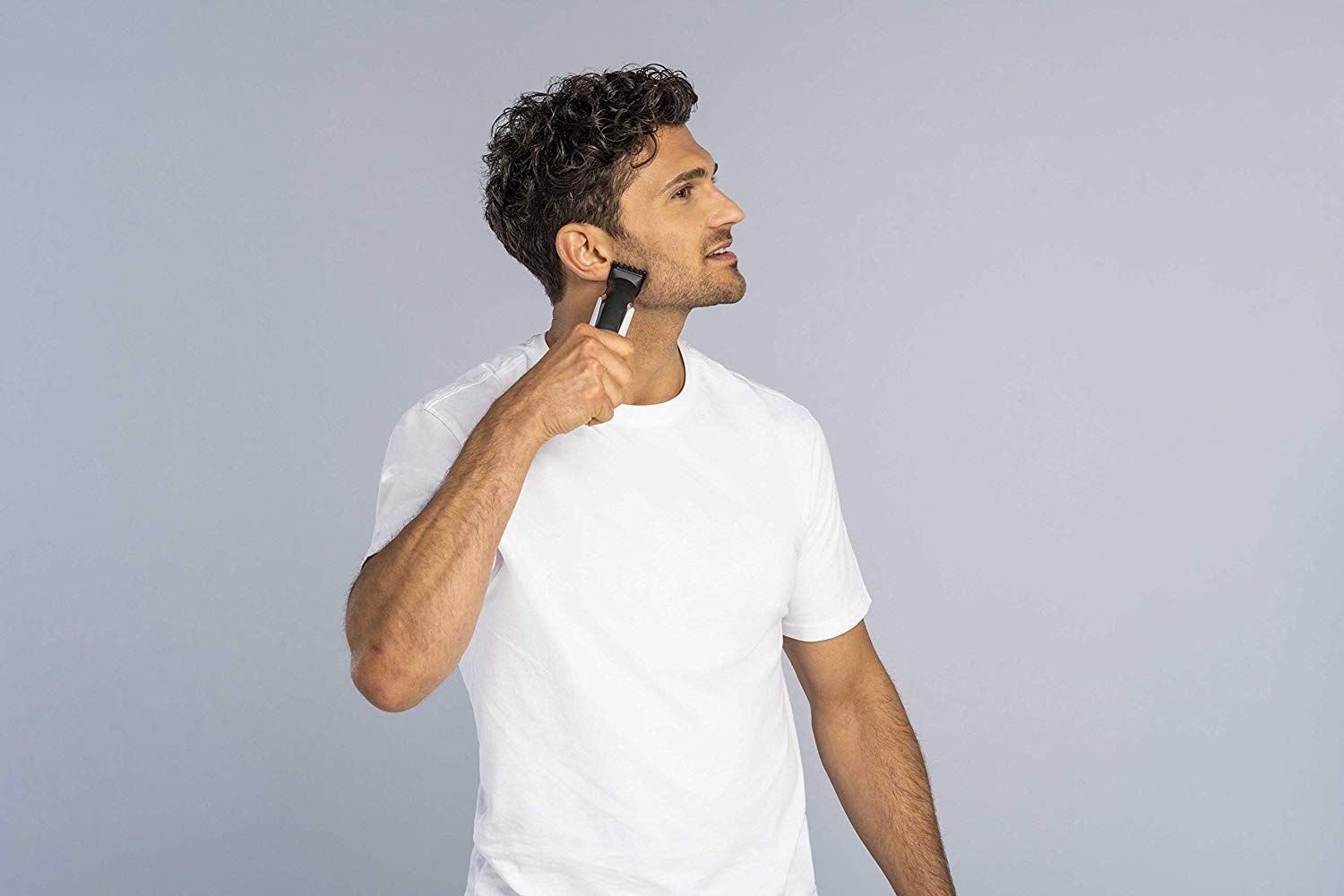 Mann rasiert sich mit einem Haarschneider. Der Mann trägt ein weißes T-Shirt. Das Gerät ist schwarz, silber und weiß.