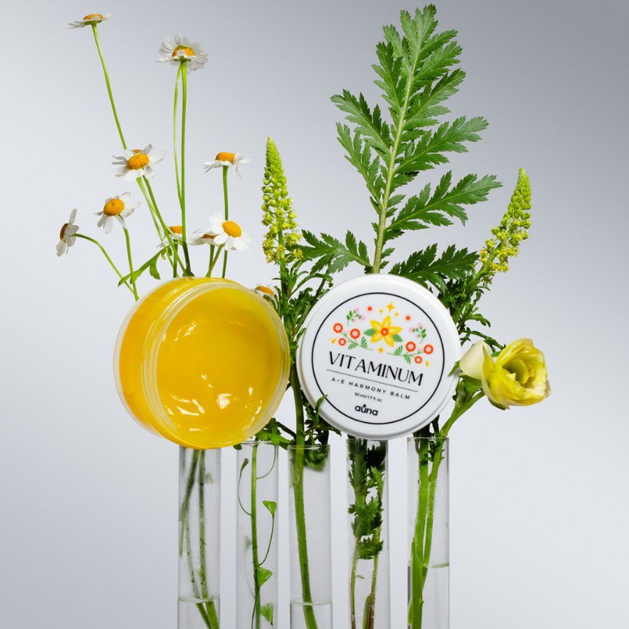 Offener Tiegel mit gelbem Balsam, neben Blumen in Vasen. Auf dem Deckel: "Vitaminum A+E Harmony Balm". Marke: auna.