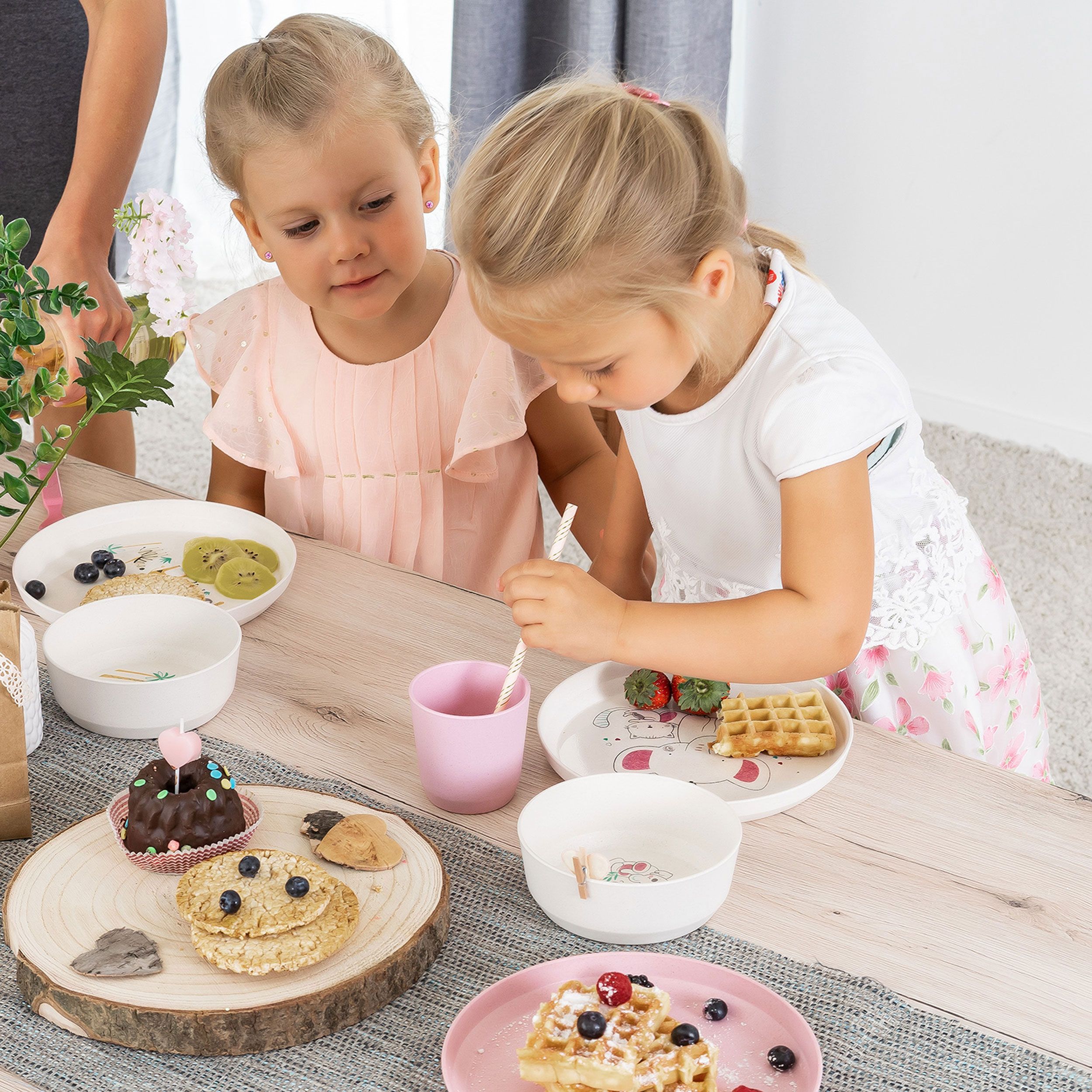 Zwei Mädchen sitzen an einem Tisch mit Kindergeschirr. Teller mit Waffeln, Beeren und Kuchen. Rosa und weiße Becher.