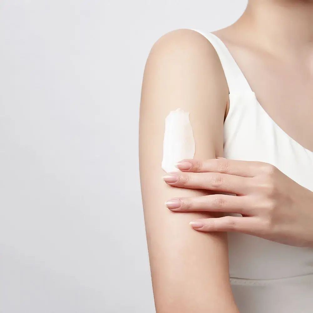 Arm mit weißer Creme bestrichen. Person trägt ein weißes Top. Heller Hintergrund.