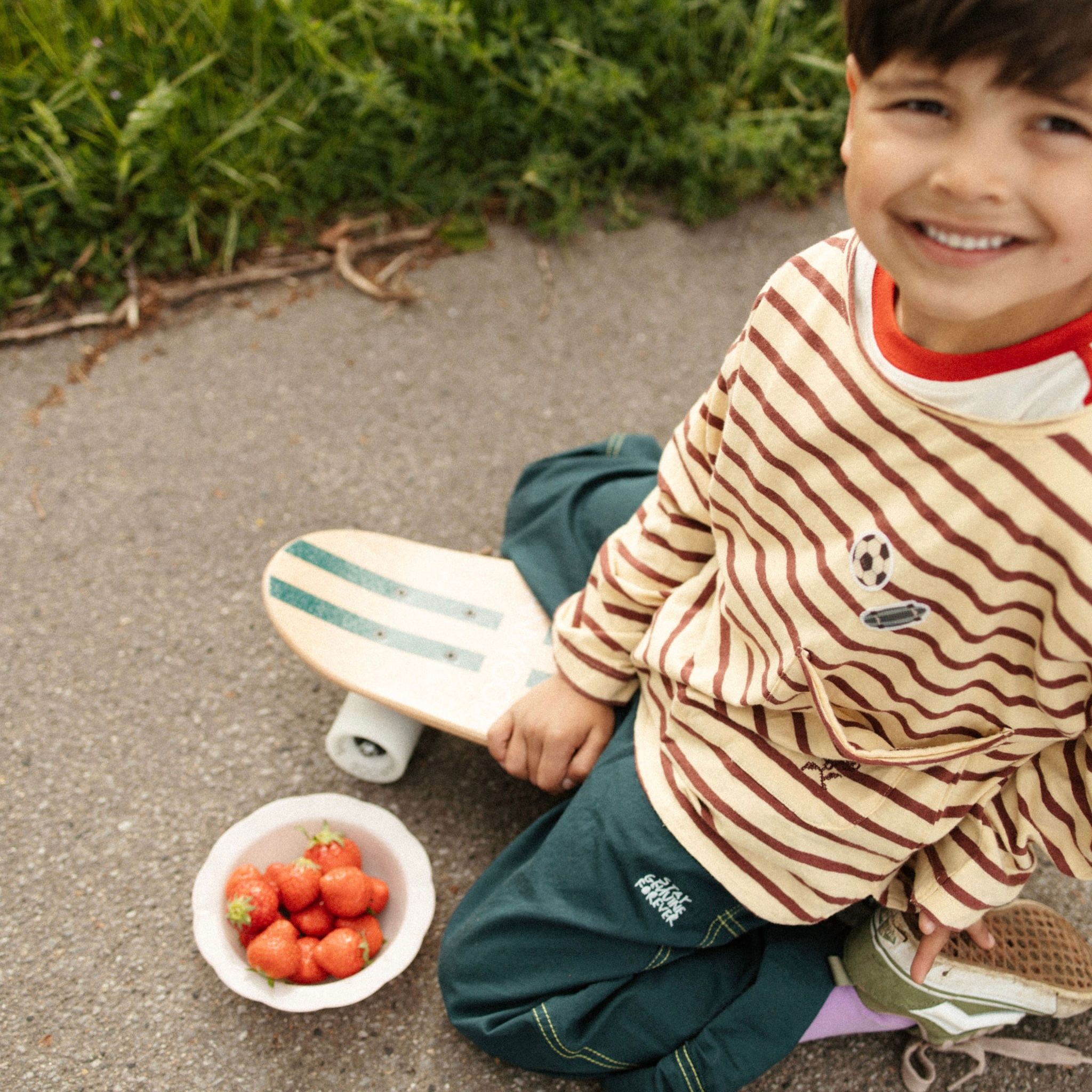Junge sitzt auf Skateboard mit Schüssel Erdbeeren, trägt gestreiftes Shirt.
