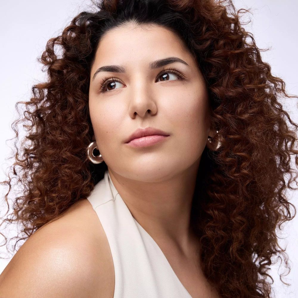 Porträt einer Frau mit lockigem Haar. Sie trägt Ohrringe und ein helles Oberteil. Blick zur Seite.