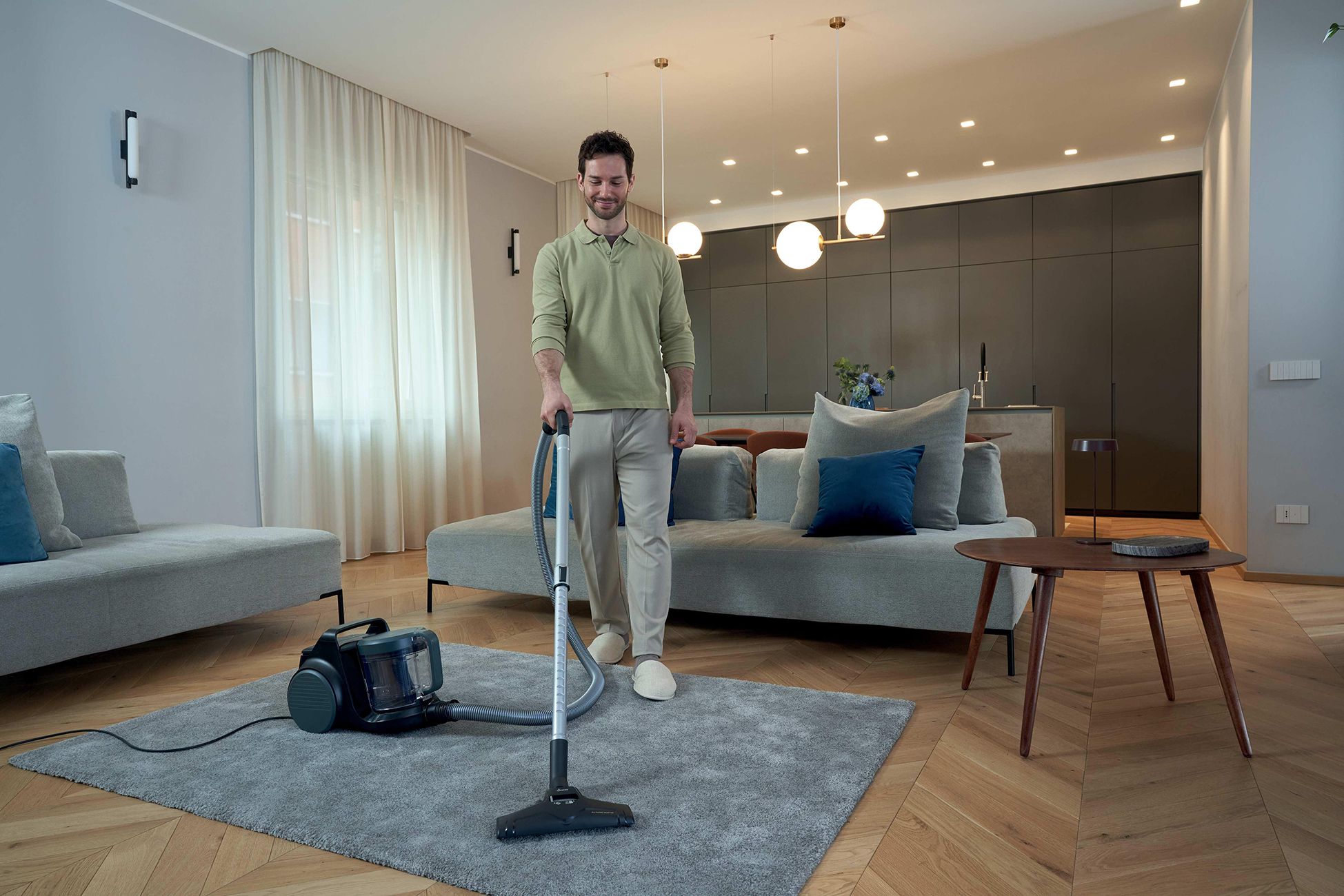 Ein Mann benutzt einen Bodenstaubsauger auf einem grauen Teppich in einem Wohnzimmer.