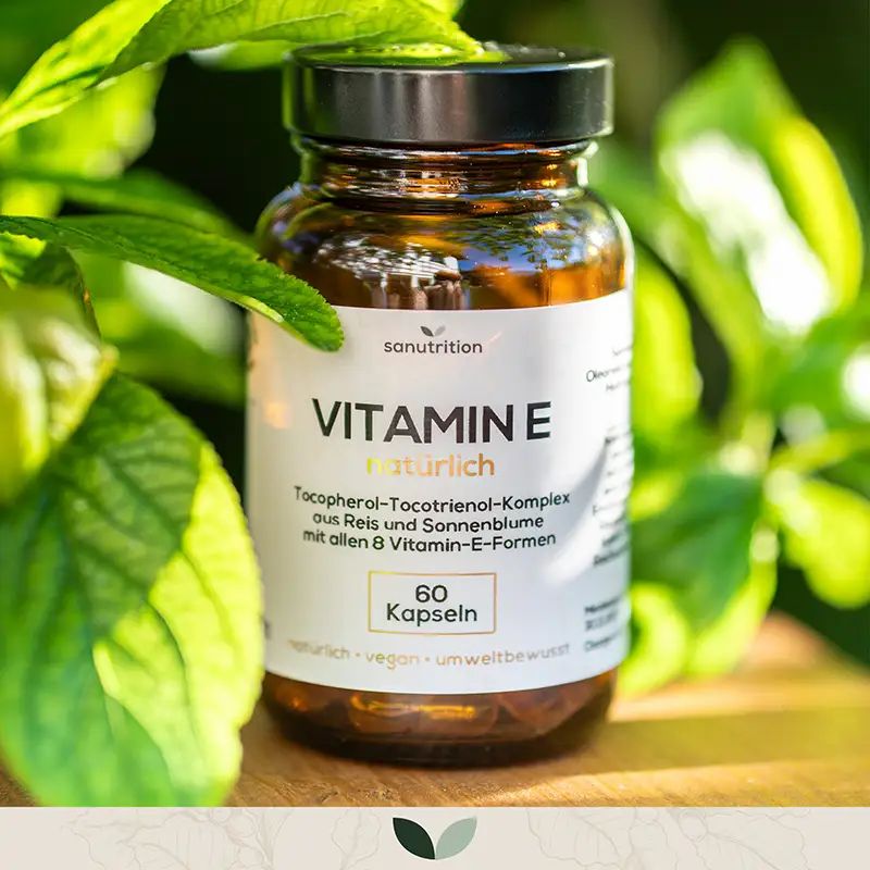 Sanutrition Vitamin E natürlich Kapseln in Braunglasflasche mit schwarzem Deckel, umgeben von grünen Blättern.