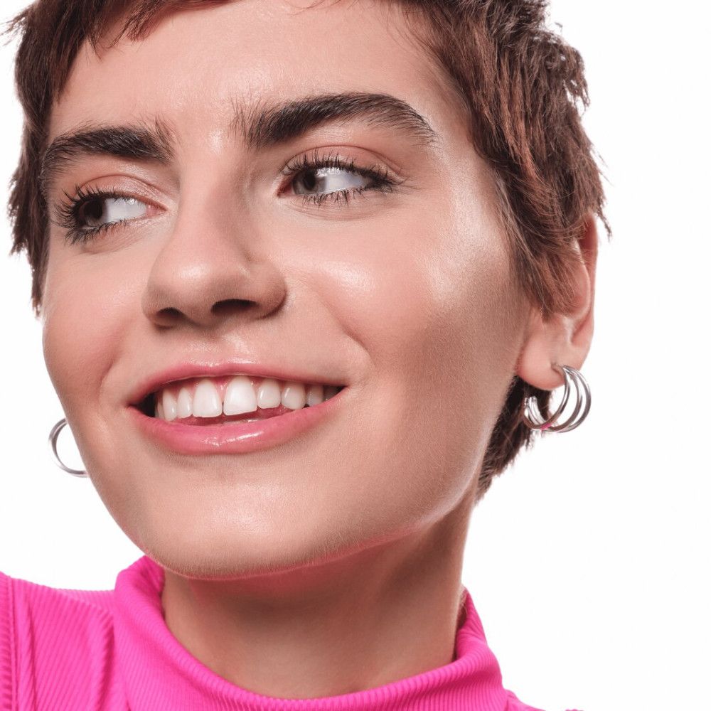 Nahaufnahme einer Frau mit kurzen Haaren, die in die Kamera lächelt. Sie trägt silberne Creolen und ein pinkes Oberteil.