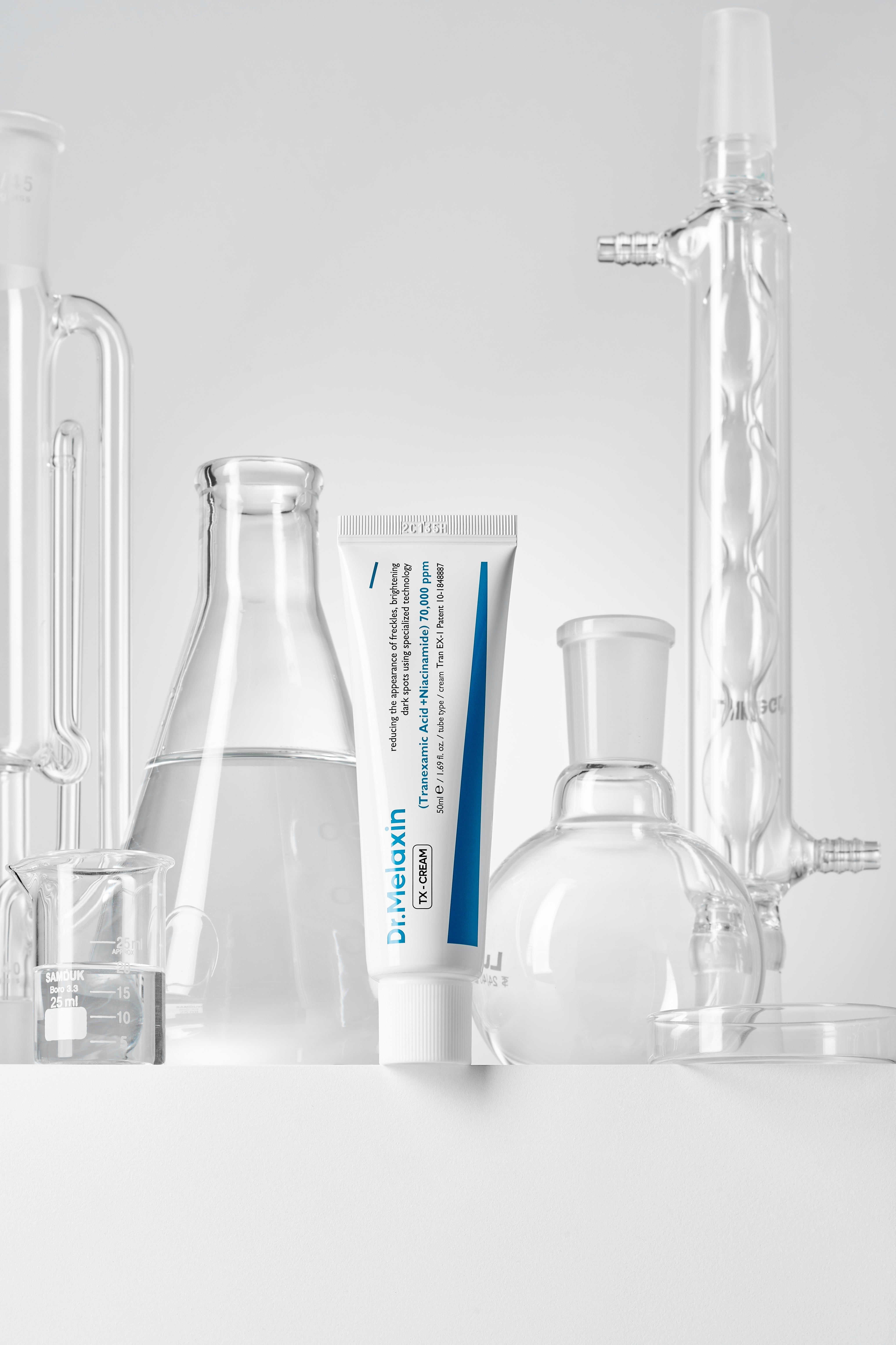 Weiße Tube mit blauer Beschriftung neben Laborgeräten. Aufschrift: Dr.Melaxin TX-CREAM. Enthält Tranexamsäure und Niacinamid.