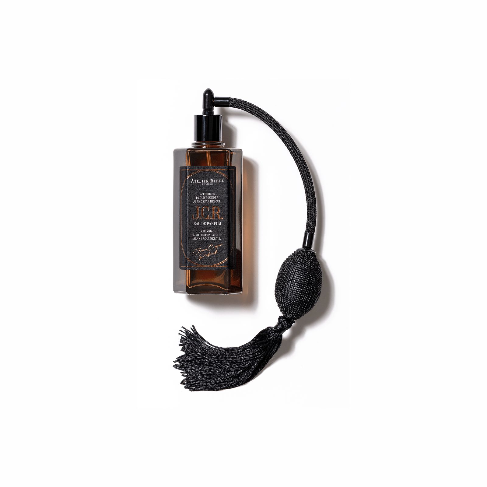 Braune Glasflasche mit schwarzem Zerstäuber und Quaste. Aufschrift: Atelier Rebul J.C.R. Eau de Parfum. Auf weißem Hintergrund.