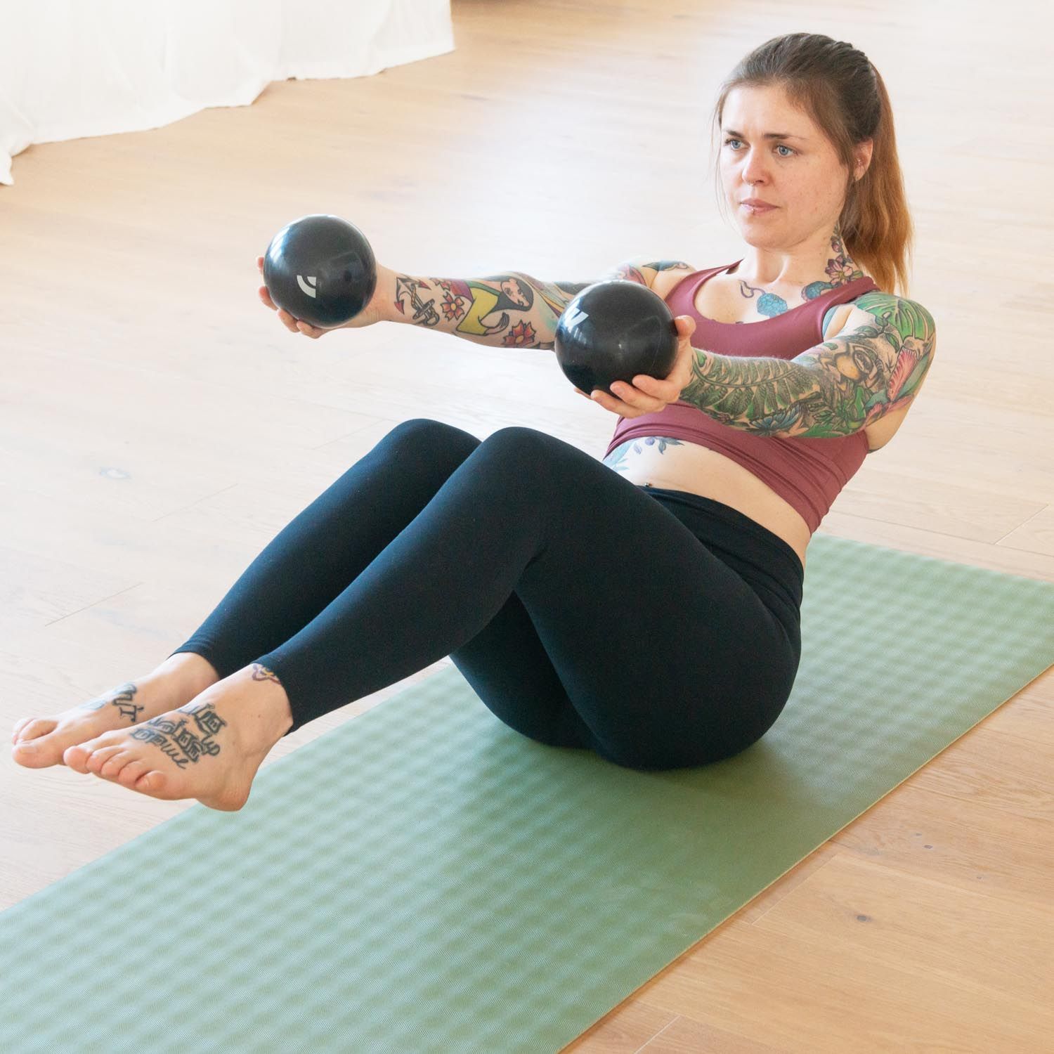 Eine Frau sitzt auf einer grünen Matte und hält zwei anthrazitfarbene Pilates-Toning Bälle. Sie trainiert.