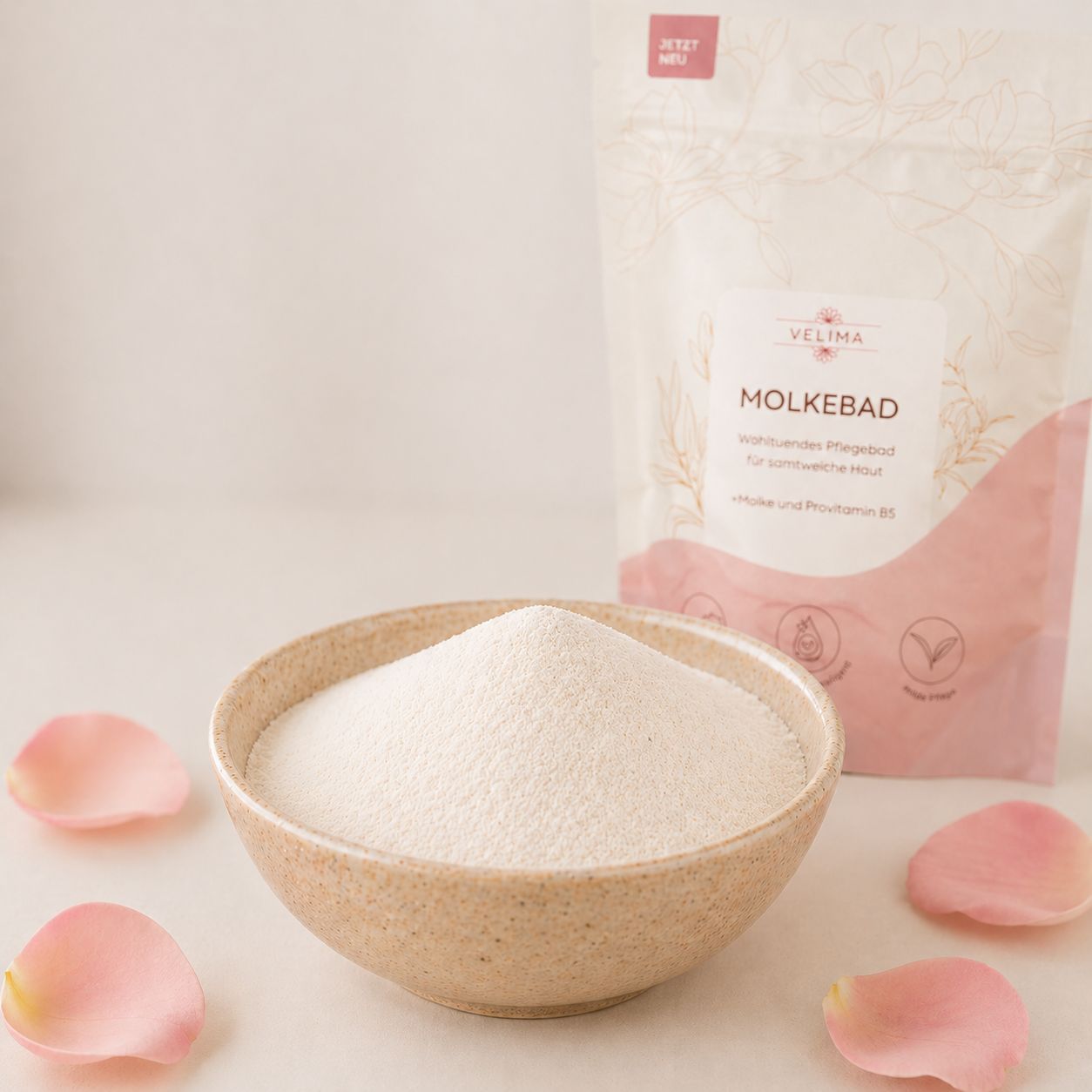 Beige Schale mit weißem Pulver, daneben eine Tüte mit Produktaufdruck. Rosenblütenblätter liegen verstreut.
