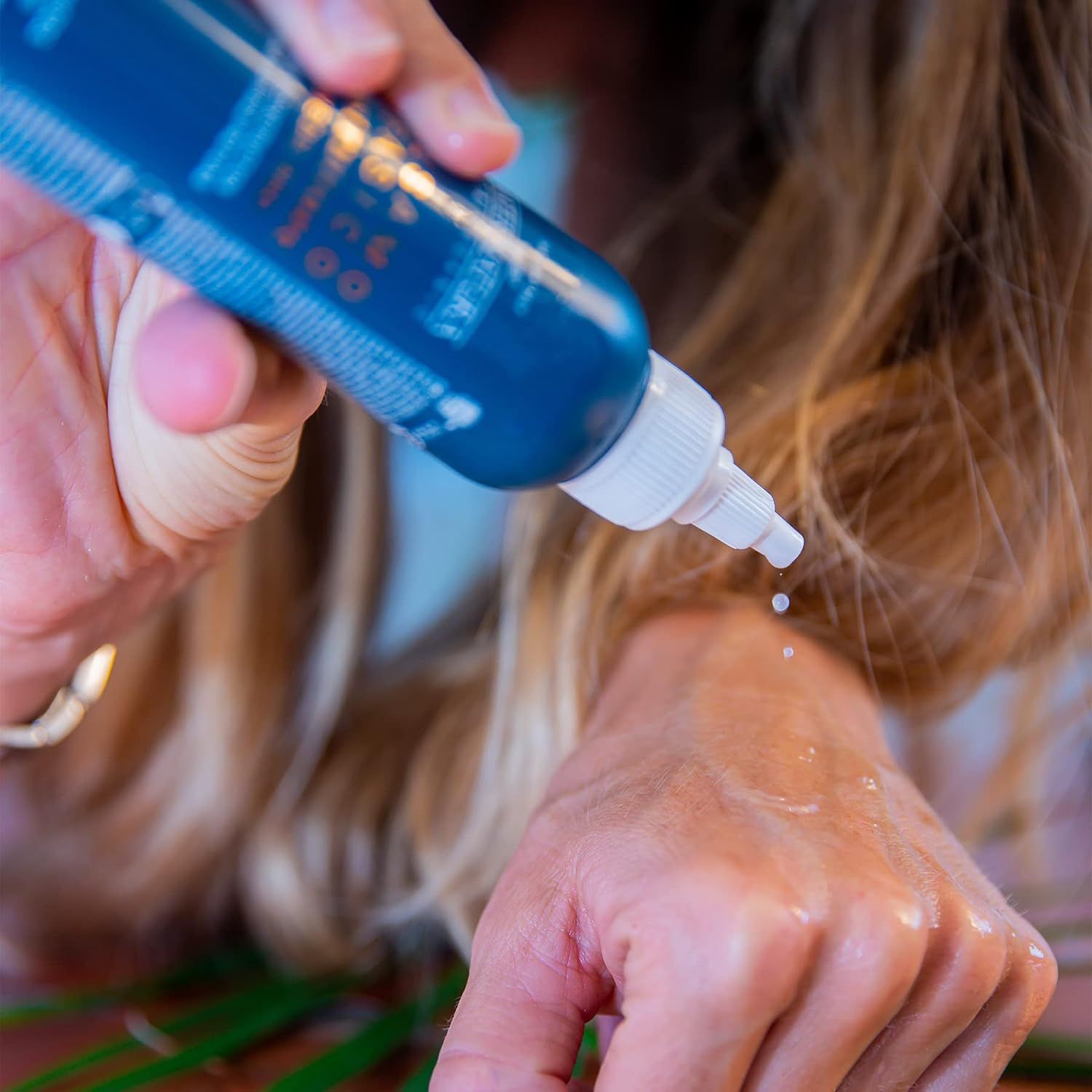 Hand drückt eine blaue Flasche mit weißem Applikator aus, Flüssigkeit tropft auf die Hand.