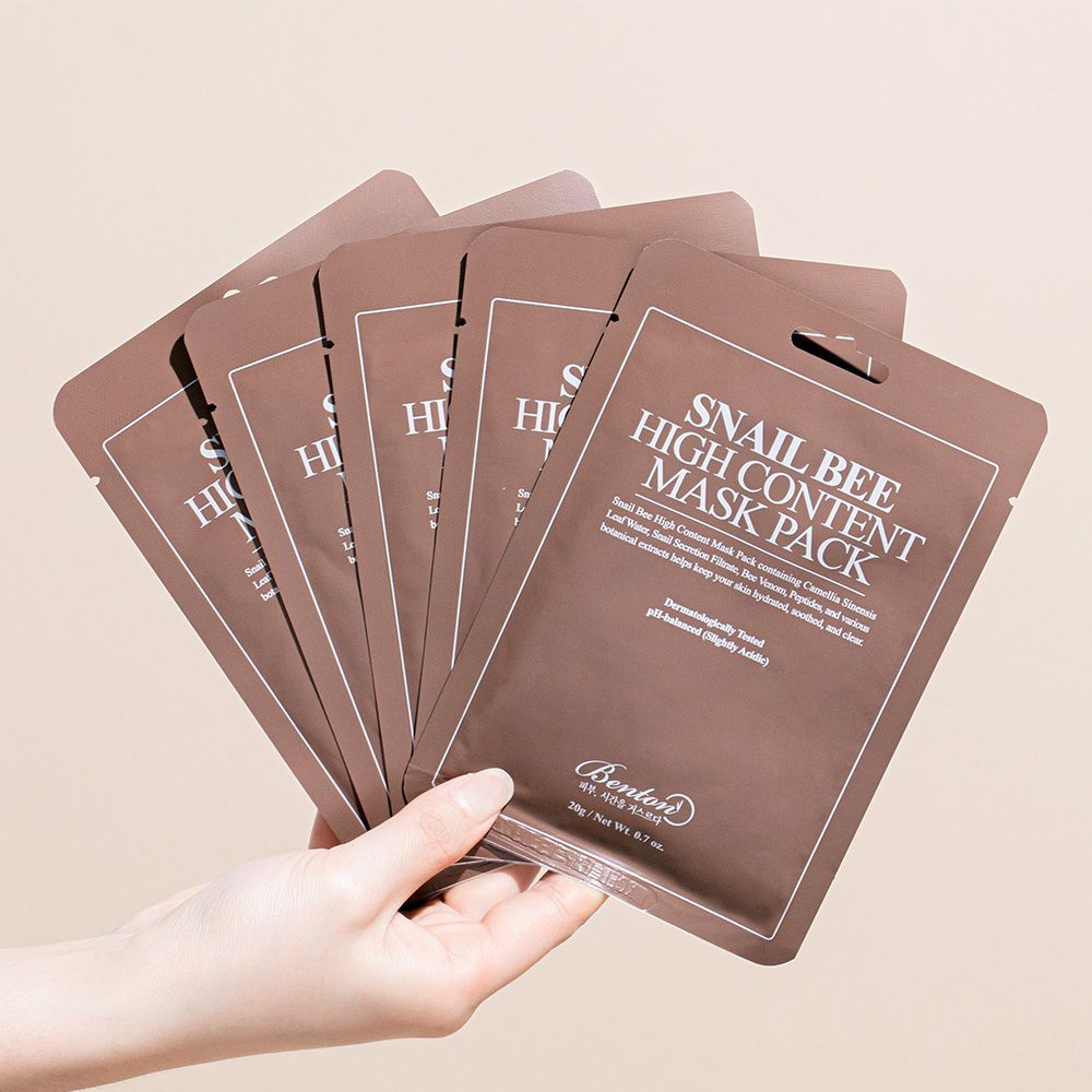 Hand hält mehrere braune Einzelverpackungen. Aufschrift: Snail Bee High Content Mask Pack. Weißer Text. Produktpräsentation.