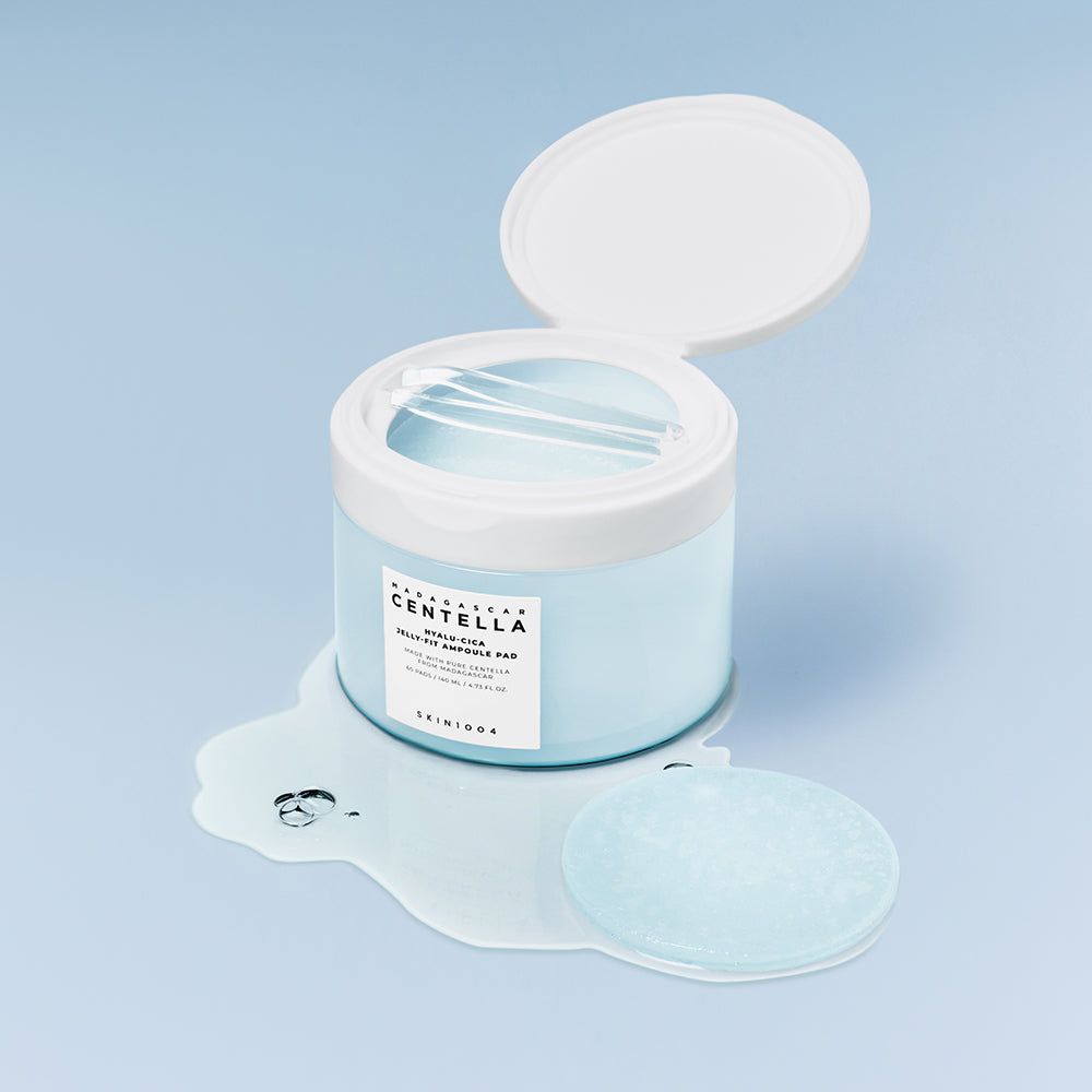 Behälter mit Deckel geöffnet, Pad und Flüssigkeit. Text: CENTELLA, HYALU-CICA JELLY-FIT AMPULE PAD, SKIN1004. Hintergrund: hellblau.