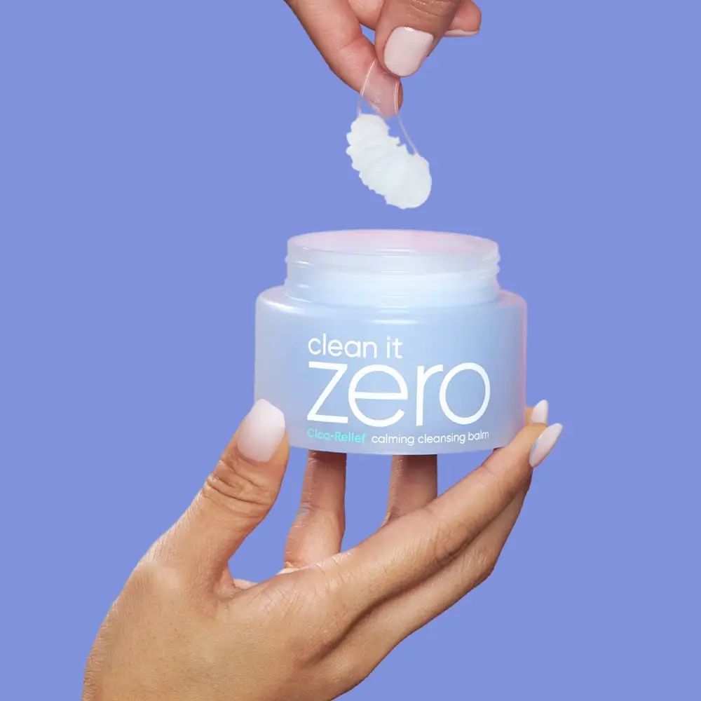 Ein blaues Glas mit 'clean it zero' und 'calming cleansing balm' Aufschrift. Eine Hand hebt eine kleine Menge des Balsams mit einem Spatel an.