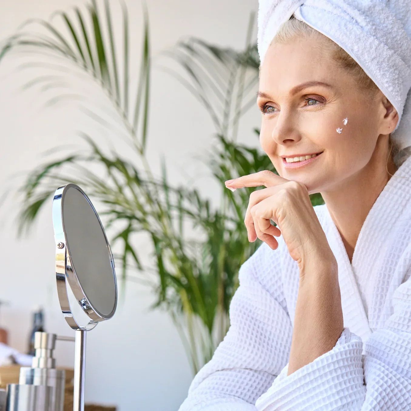 Frau mit Handtuch und Bademantel vor Spiegel. Auf der Haut: Creme-Punkte.